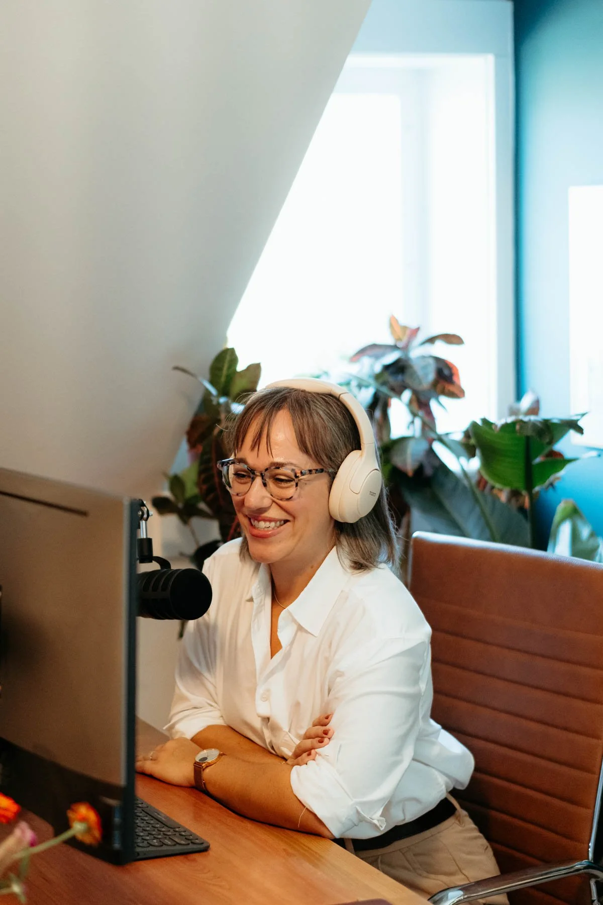 A woman with glasses and a white blouse, smiling while wearing headphones and sitting at a wooden desk in front of a computer monitor. Behind her are large green plants and a window with light coming through.