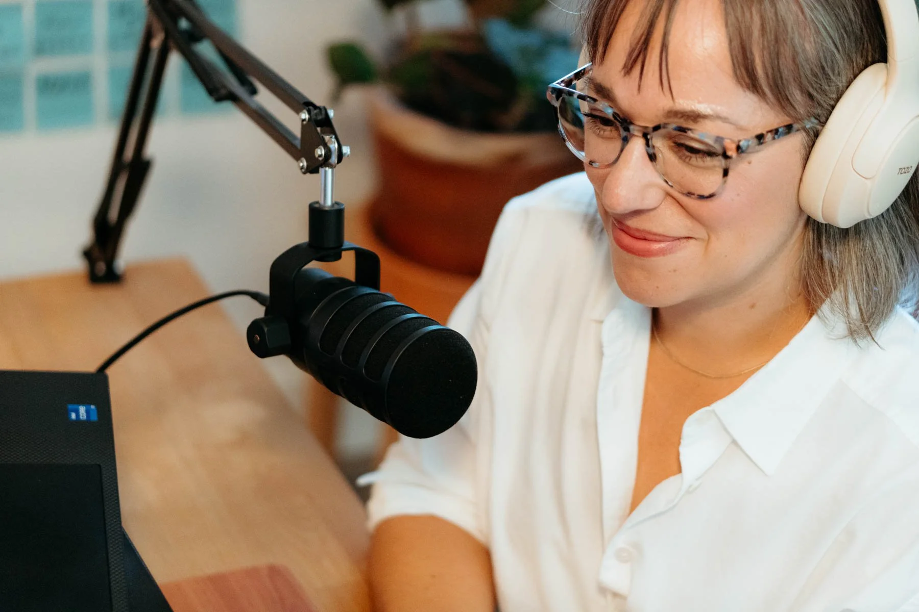 A woman with short brown hair, glasses, and headphones speaking into a black microphone during a recording session.