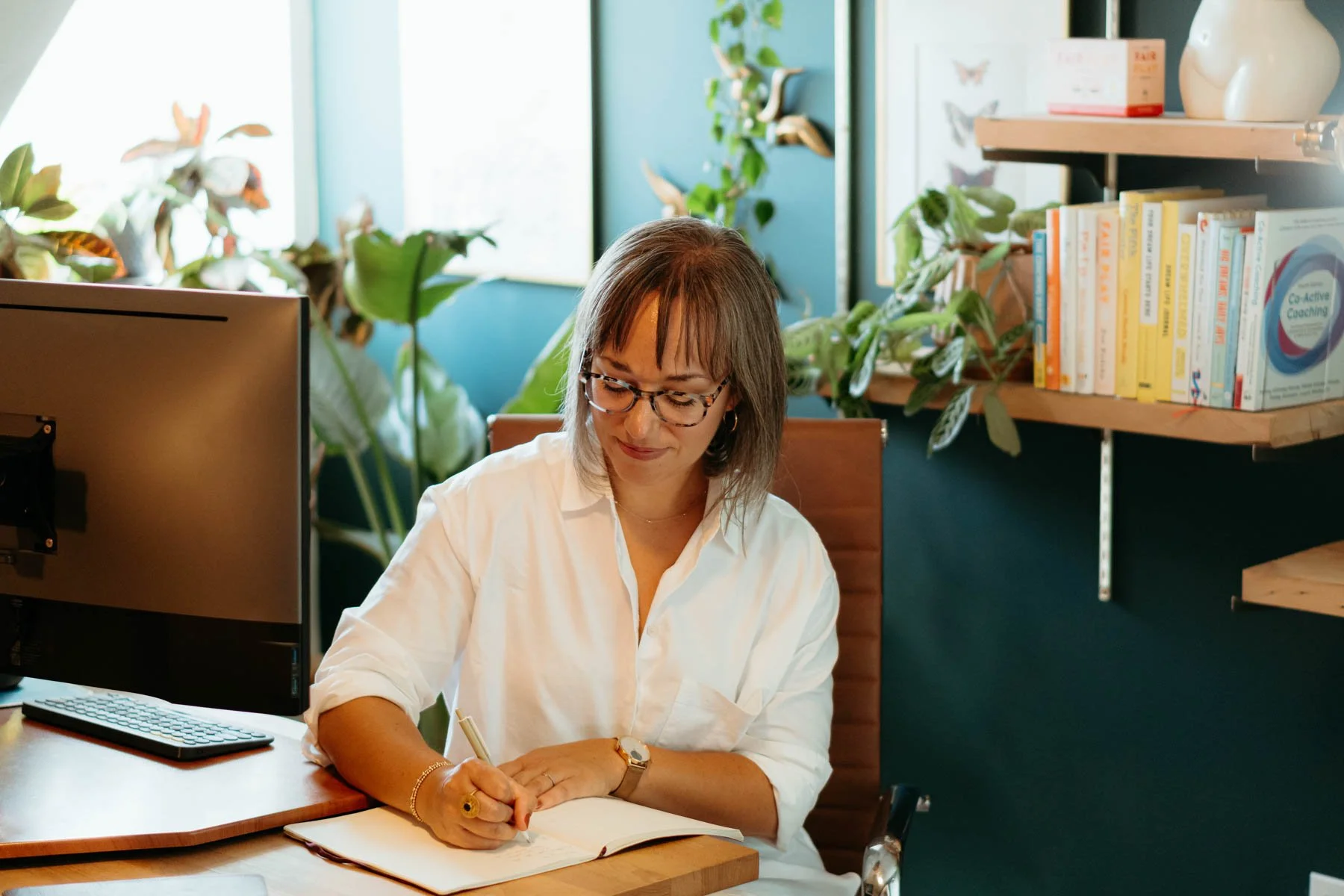 A woman with glasses and shoulder-length brown hair, dressed in a white blouse, sitting at a desk in a bright, plant-filled office. She is writing in a notebook with a pen, with a computer monitor on her left and books on shelves behind her.