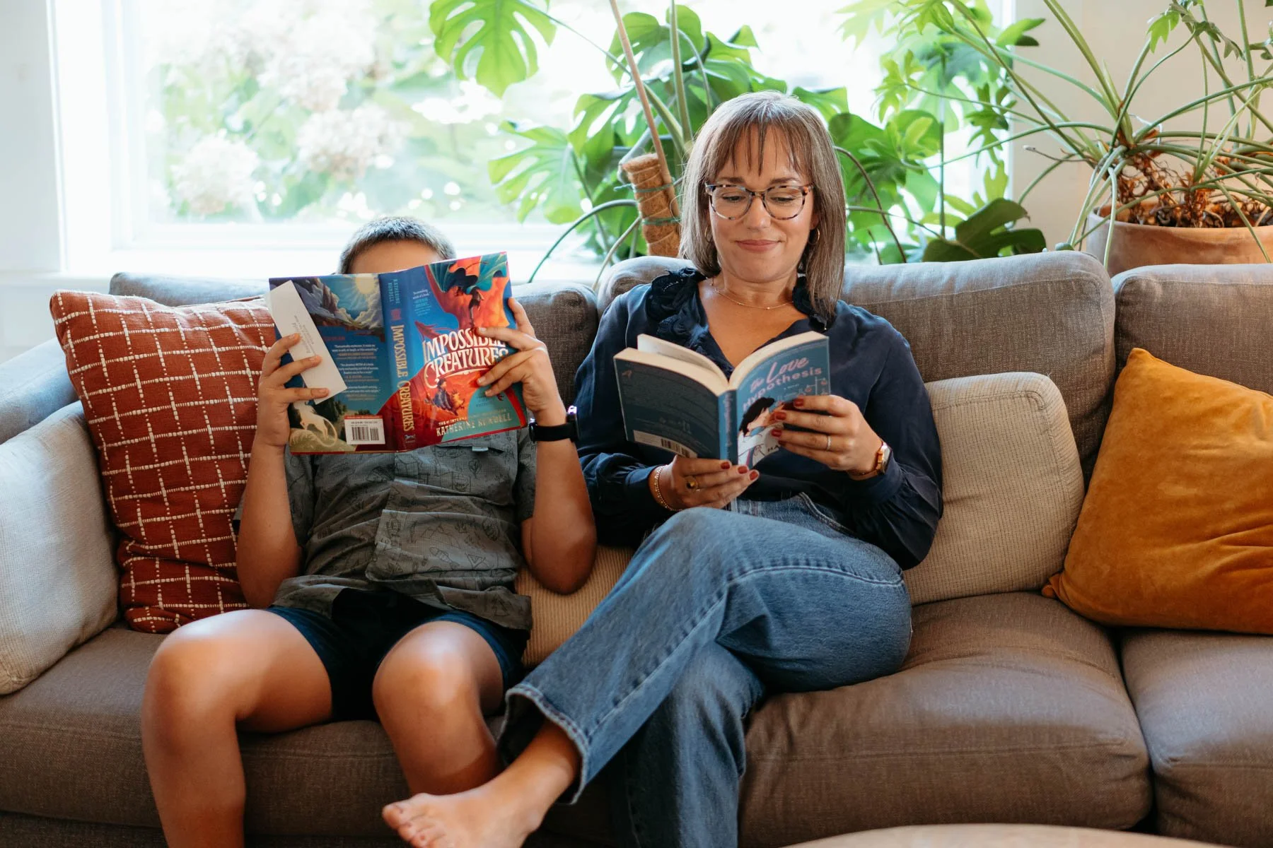 A woman and a young boy sitting on a beige sofa, reading books. The woman is smiling, wearing glasses, a dark top, and jeans. The boy is holding a colorful book and appears to be lying back. There are patterned and solid colored pillows around them, with large green plants and a window in the background.