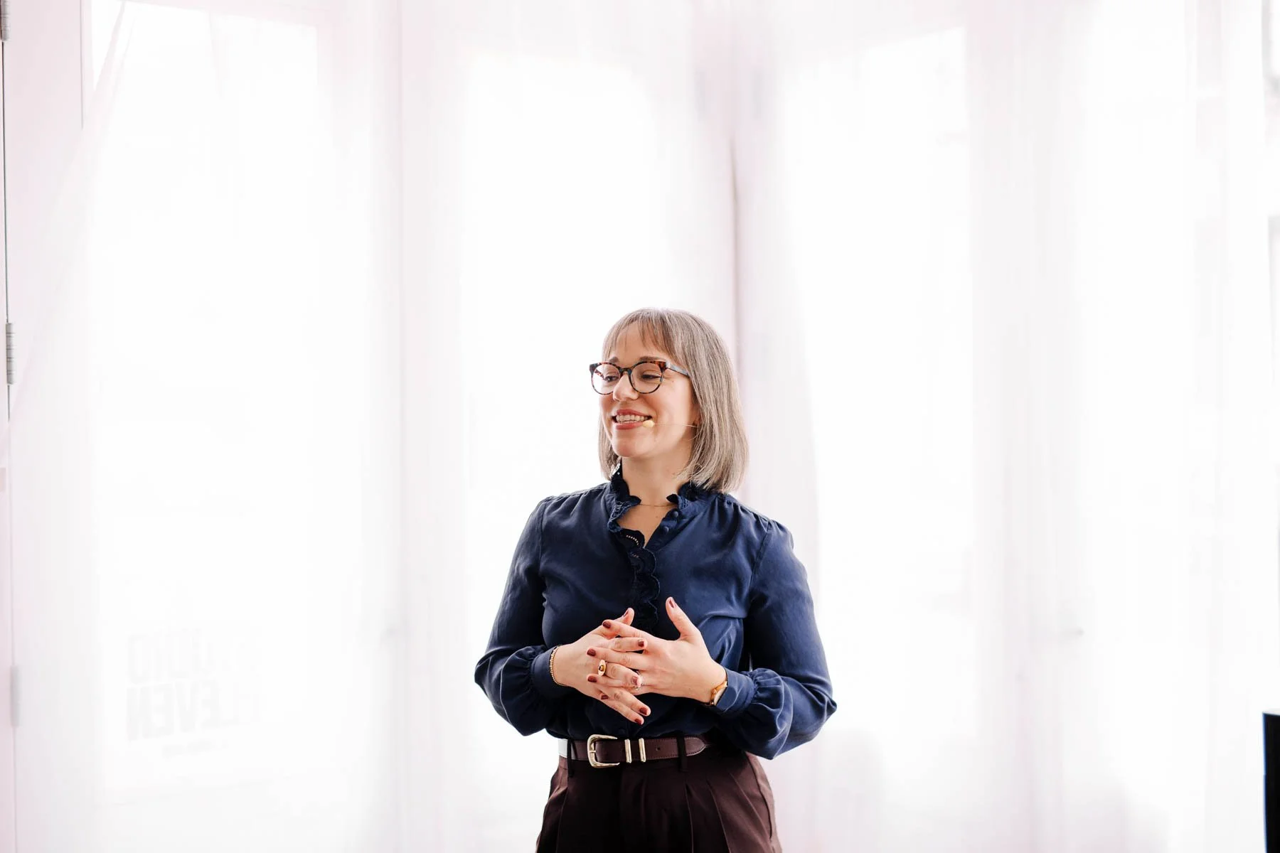 Woman smiling, wearing glasses and a dark blue blouse, standing in front of white curtains.