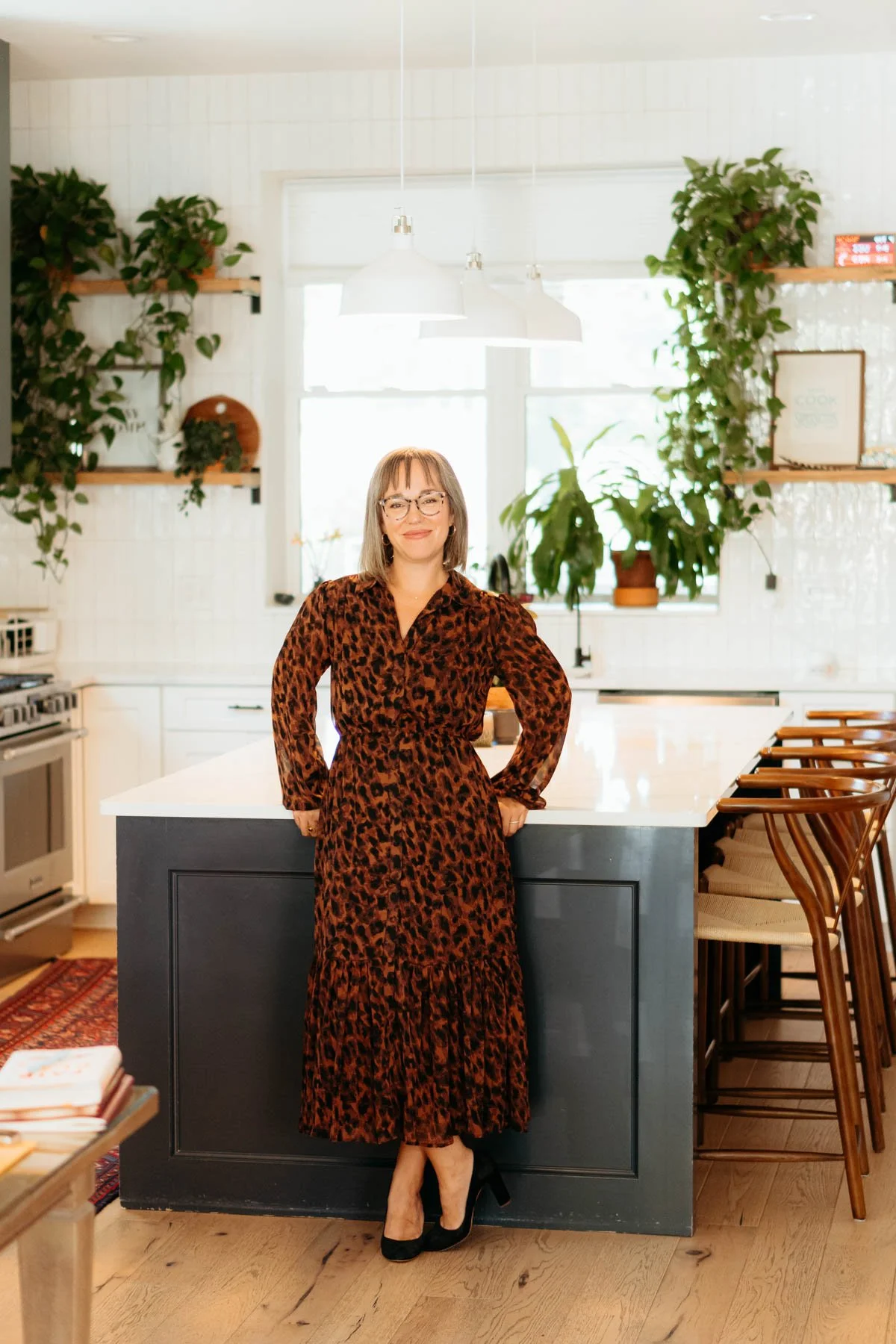 A woman in a long animal print dress standing in a modern kitchen with green plants, white cabinets, and a white countertop.
