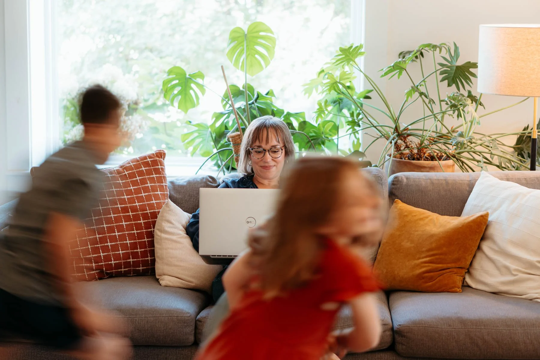 Woman sitting on a sofa using a laptop, with children blurred moving around her, in a bright room with large windows, green plants, and cushions.