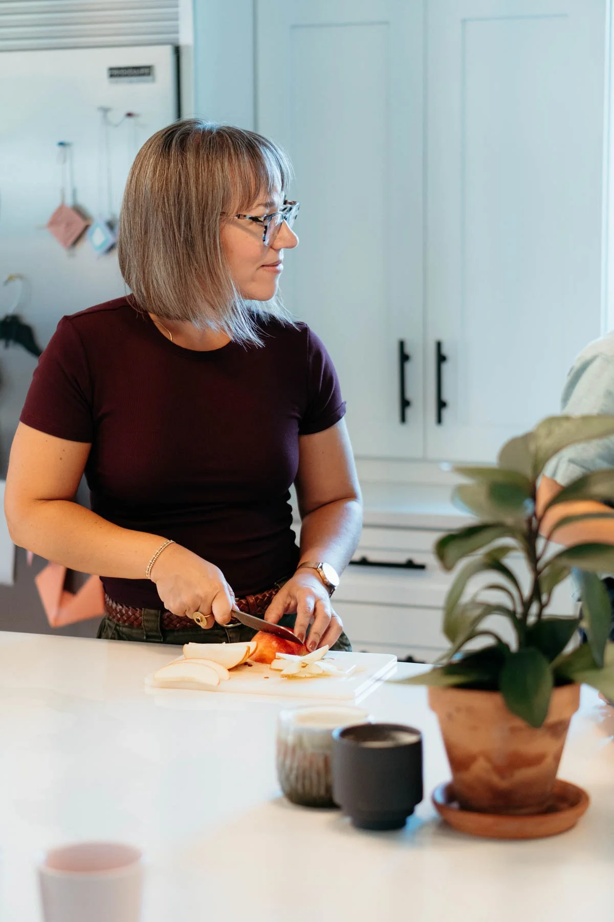 Woman with glasses and short hair slicing an apple on a kitchen counter, with a potted plant in the foreground.