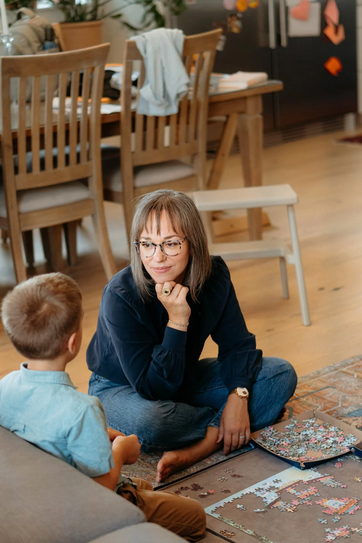 A woman with glasses and a black shirt sits on the floor, looking at a young boy with light brown hair, who is sitting on a couch. They are working on assembling a colorful puzzle on the floor in a cozy room with wooden furniture and a refrigerator in the background.