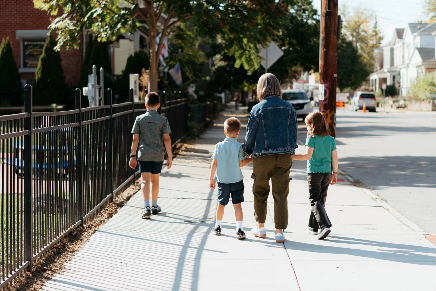 A woman walking with four children on a sunny sidewalk in a residential neighborhood. There are houses, trees, parked cars, and a utility pole along the street.