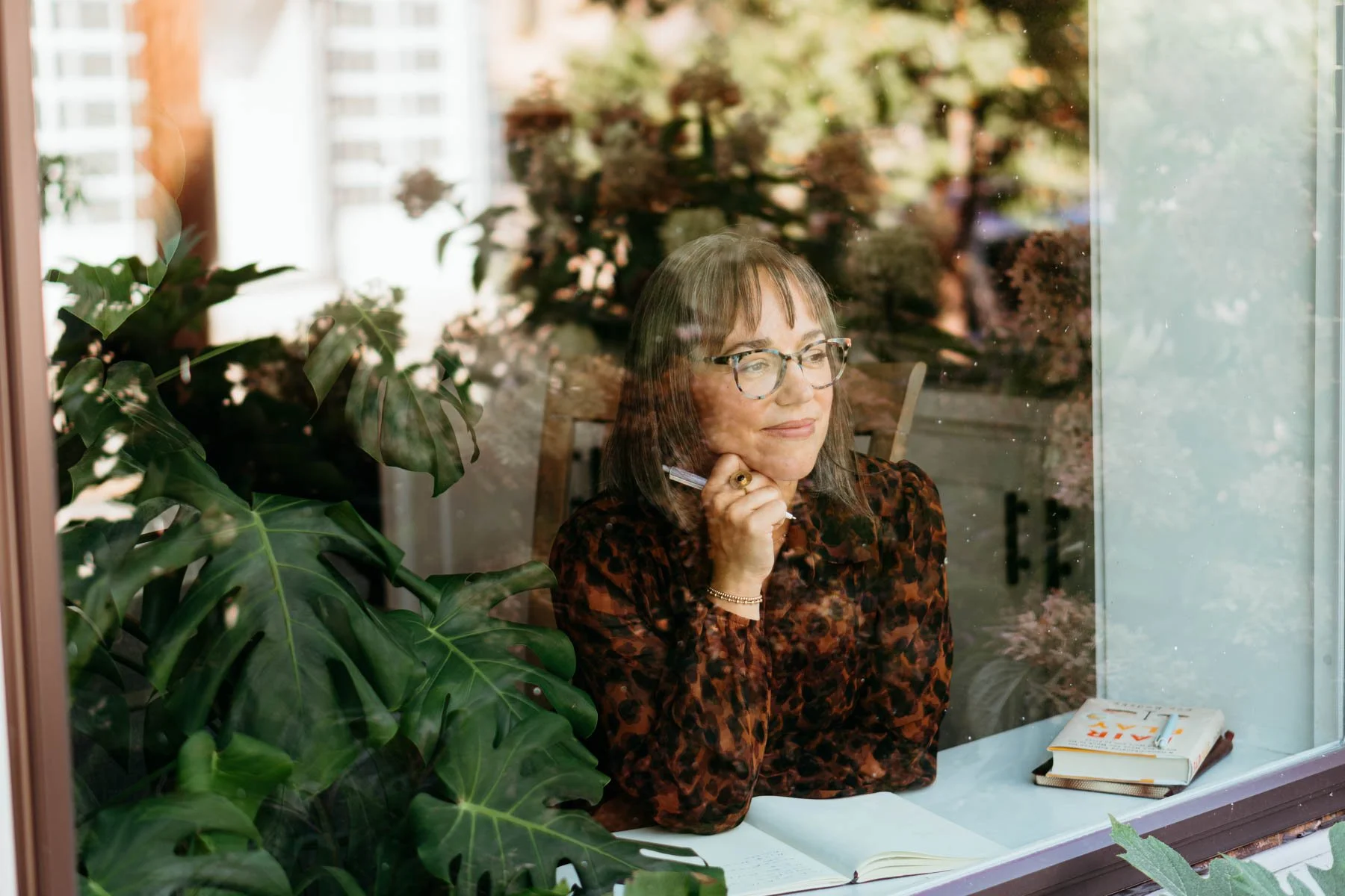 A woman with glasses and brown hair sitting at a desk behind a glass window, with green plants surrounding her, holding a pen near her chin, and looking thoughtfully outside, with books and papers on the desk.