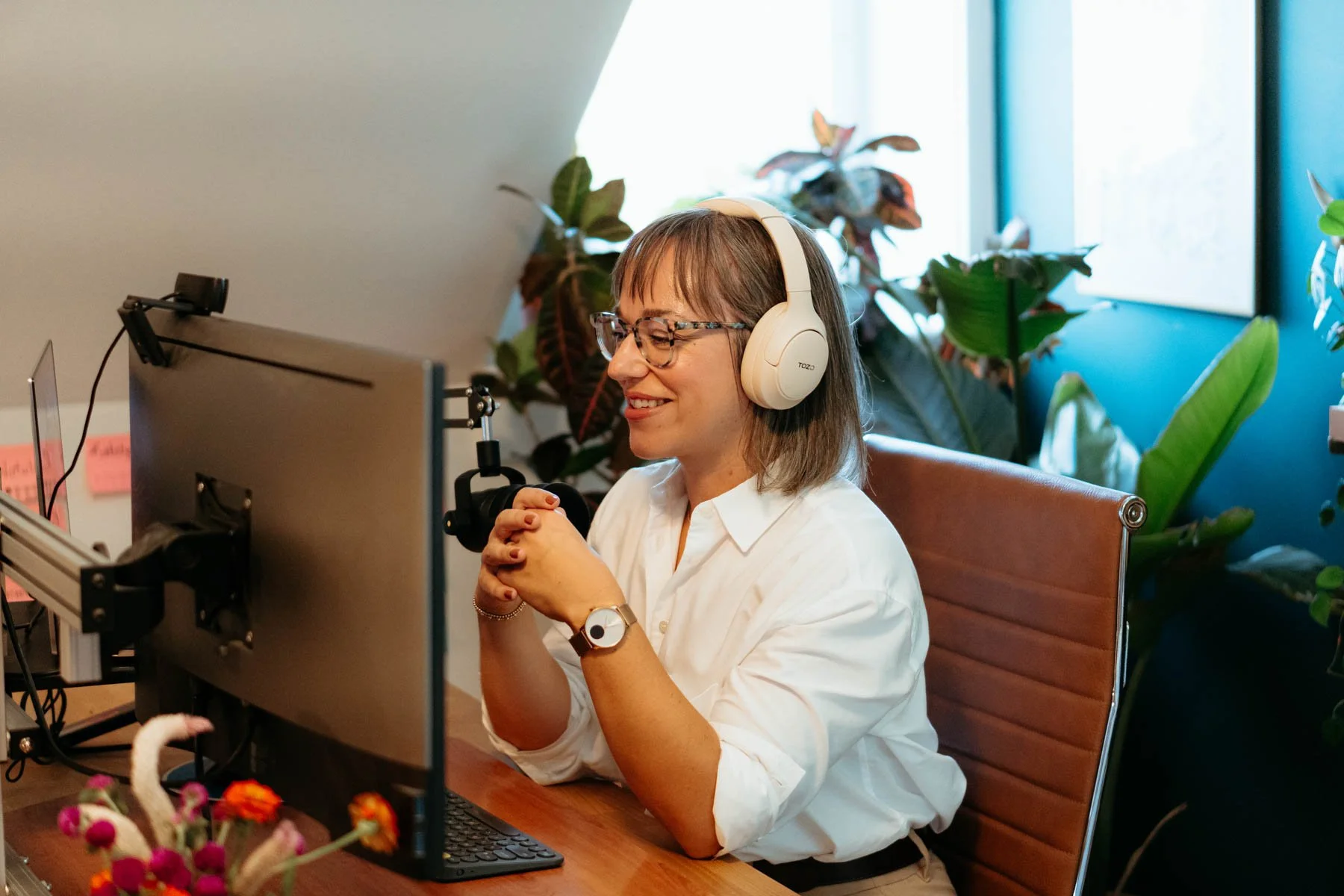 A woman with shoulder-length brown hair wearing glasses and a white blouse is sitting at a desk in front of a computer, smiling with her hands clasped. She has on a watch and a bracelet, and is wearing large white headphones. Behind her are green plants and a window with blue walls, indicating a bright, modern office environment.