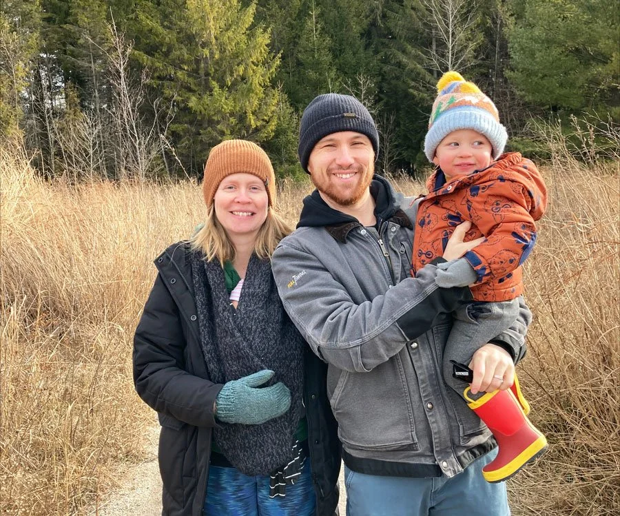 A family of three outdoors in a field of tall grass with trees in the background, dressed in warm clothing, smiling at the camera. The man is holding a child, and a woman stands beside them.