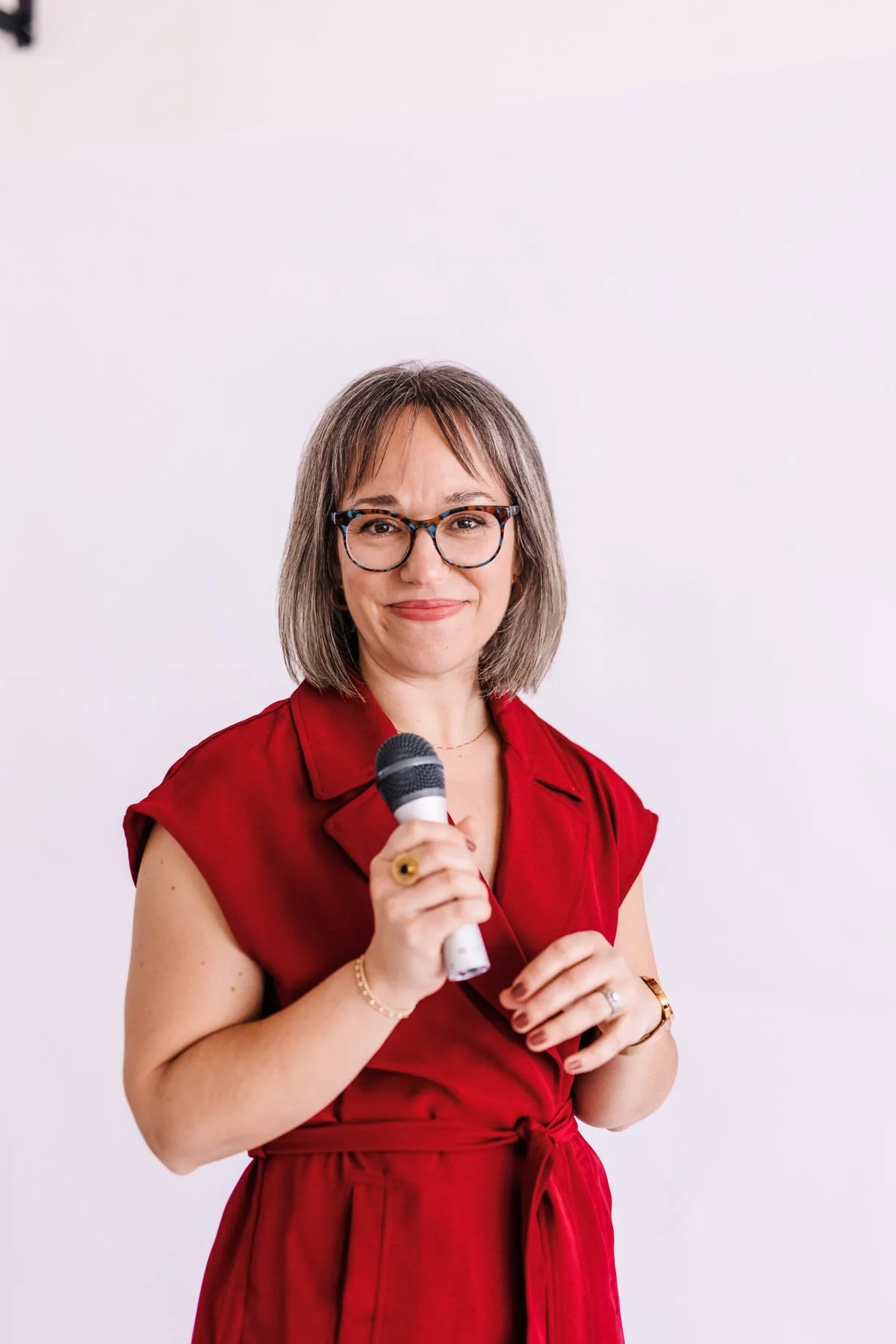 A woman with glasses and shoulder-length hair smiling and holding a microphone, wearing a red dress, standing against a plain white background.