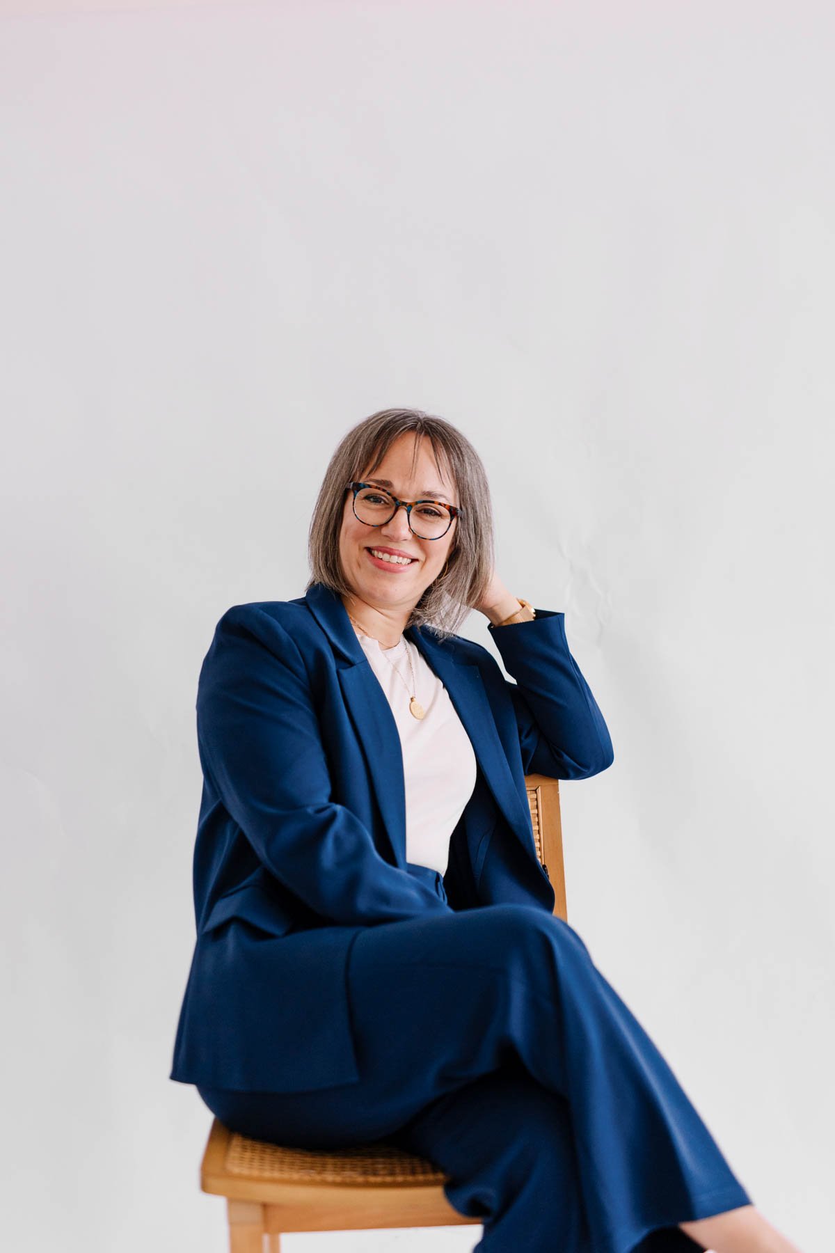 A woman sitting on a wooden chair, smiling at the camera. She is wearing a navy blue blazer and pants, a white top, glasses, and gold jewelry, with a plain light-colored background.