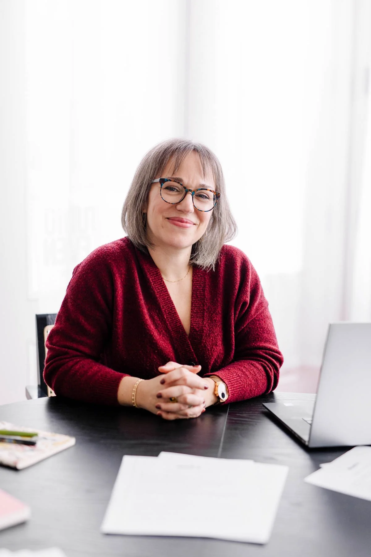 A woman with shoulder-length gray hair, wearing glasses and a red sweater, smiling and sitting at a desk in an office setting.