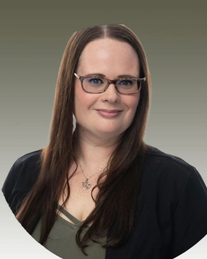 A woman with long brown hair wearing glasses, a black jacket, a green top, and a necklace, smiling against a gray gradient background.