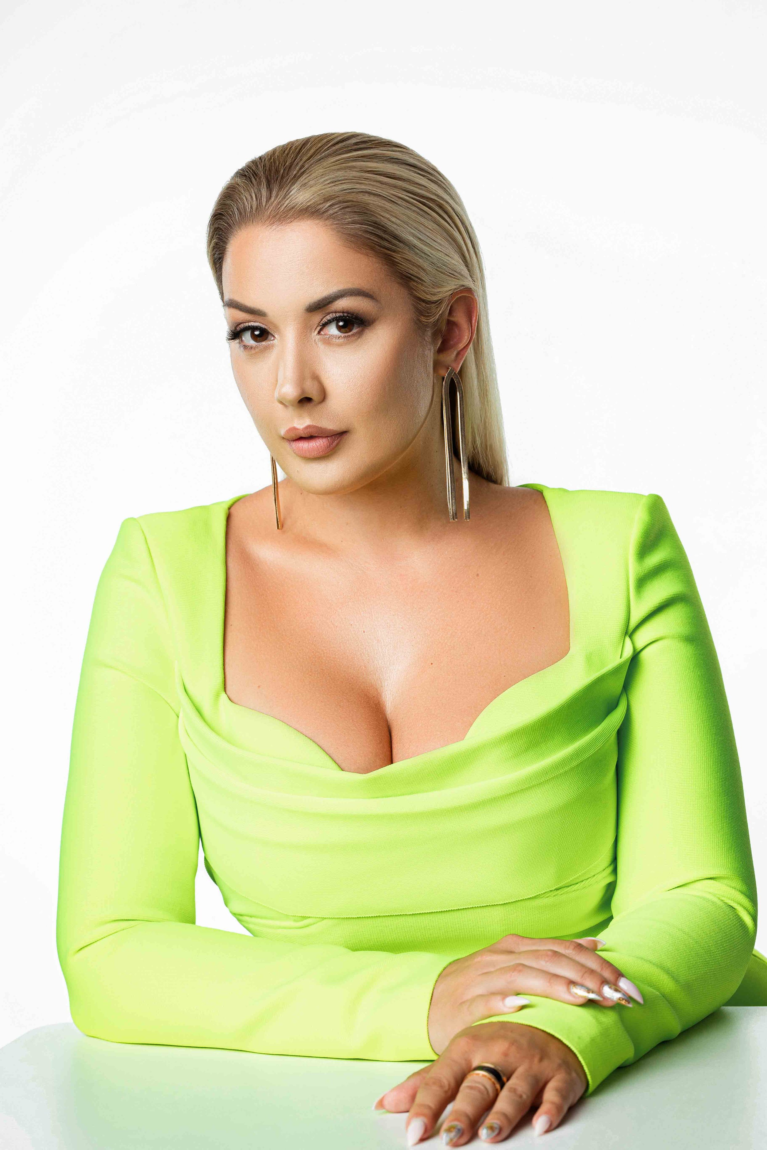 A woman with blonde hair wearing a neon green dress and large earrings, sitting with her hands on a white surface, facing the camera against a plain white background.