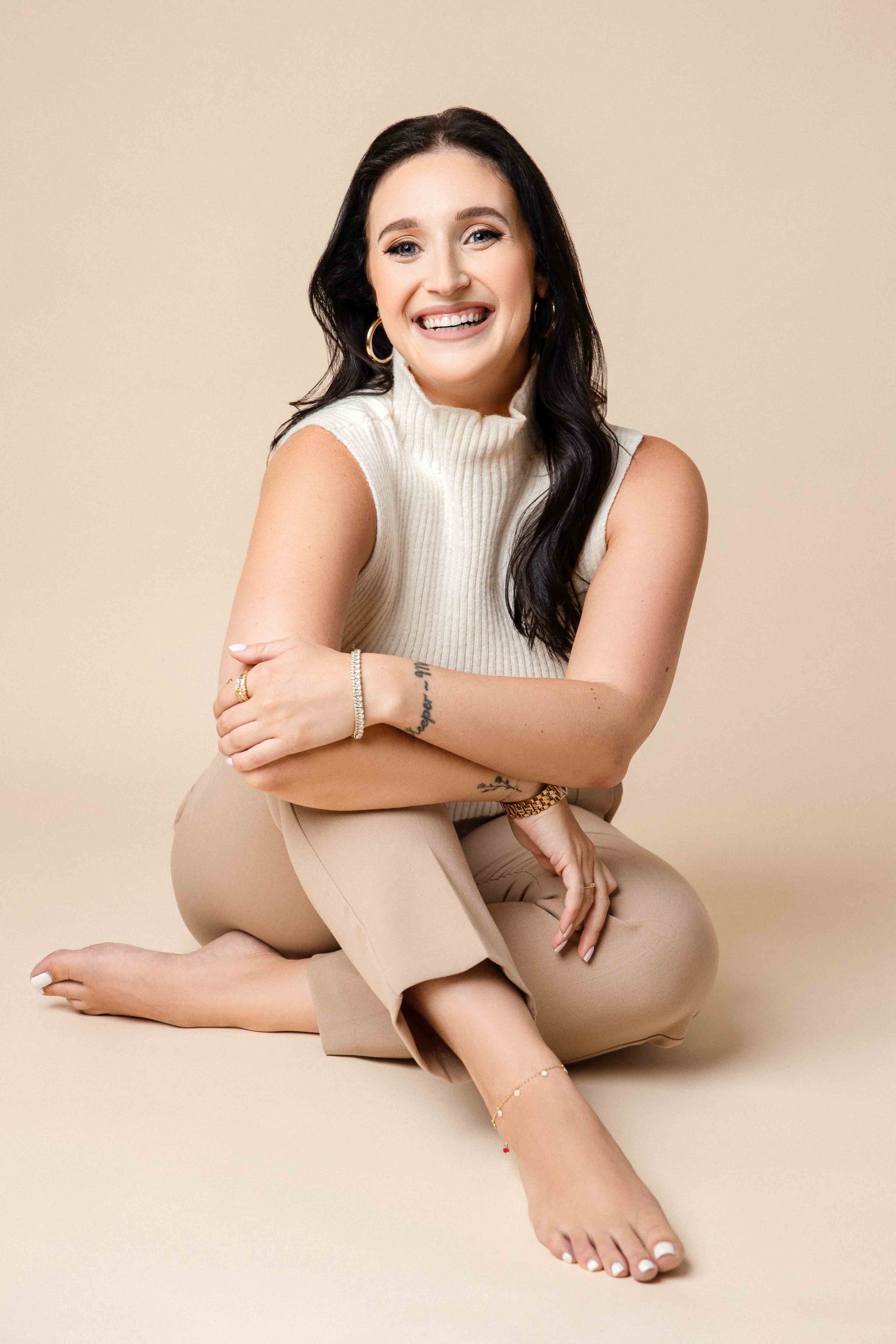 A woman with dark hair, wearing a cream sleeveless turtleneck, beige pants, and jewelry, sitting on the floor with a neutral background, smiling with arms crossed.