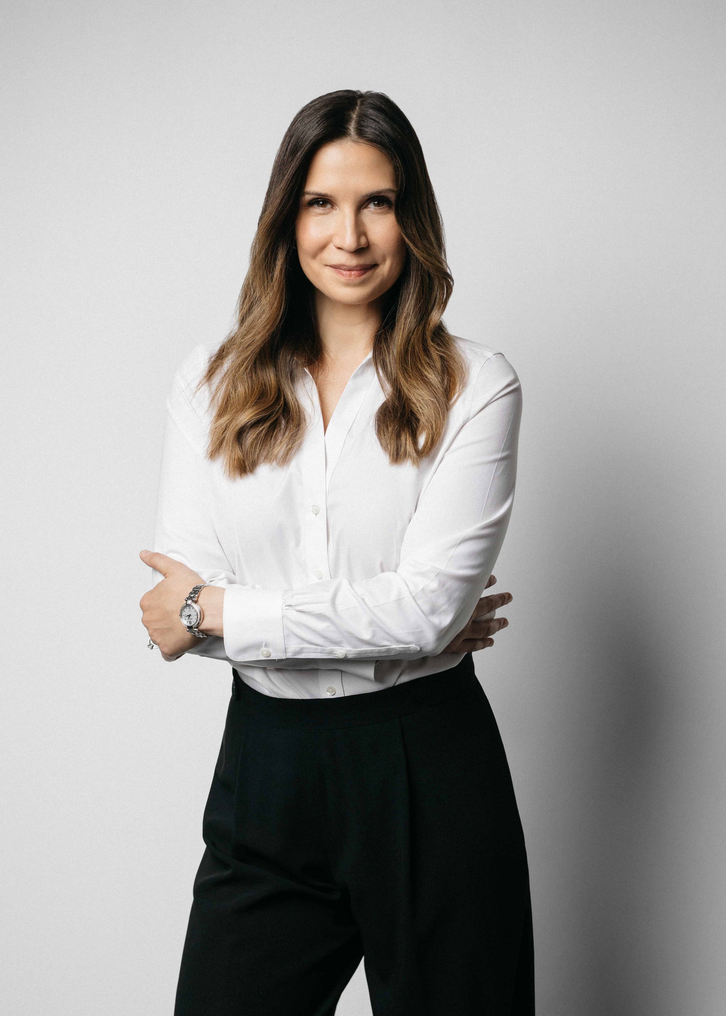 Portrait of a woman with long brown hair wearing a white button-up shirt and black pants, standing with arms crossed against a plain light gray background.