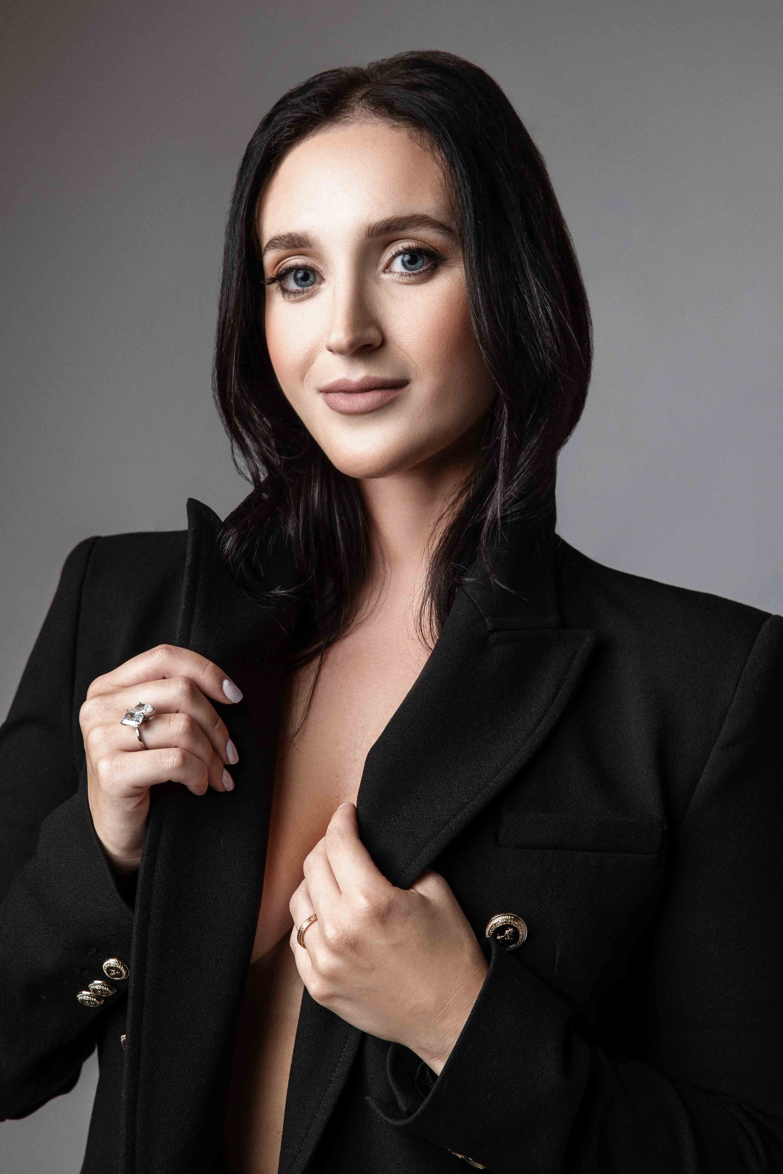 A woman with long dark hair, blue eyes, and fair skin wearing a black blazer, posing against a gray background.