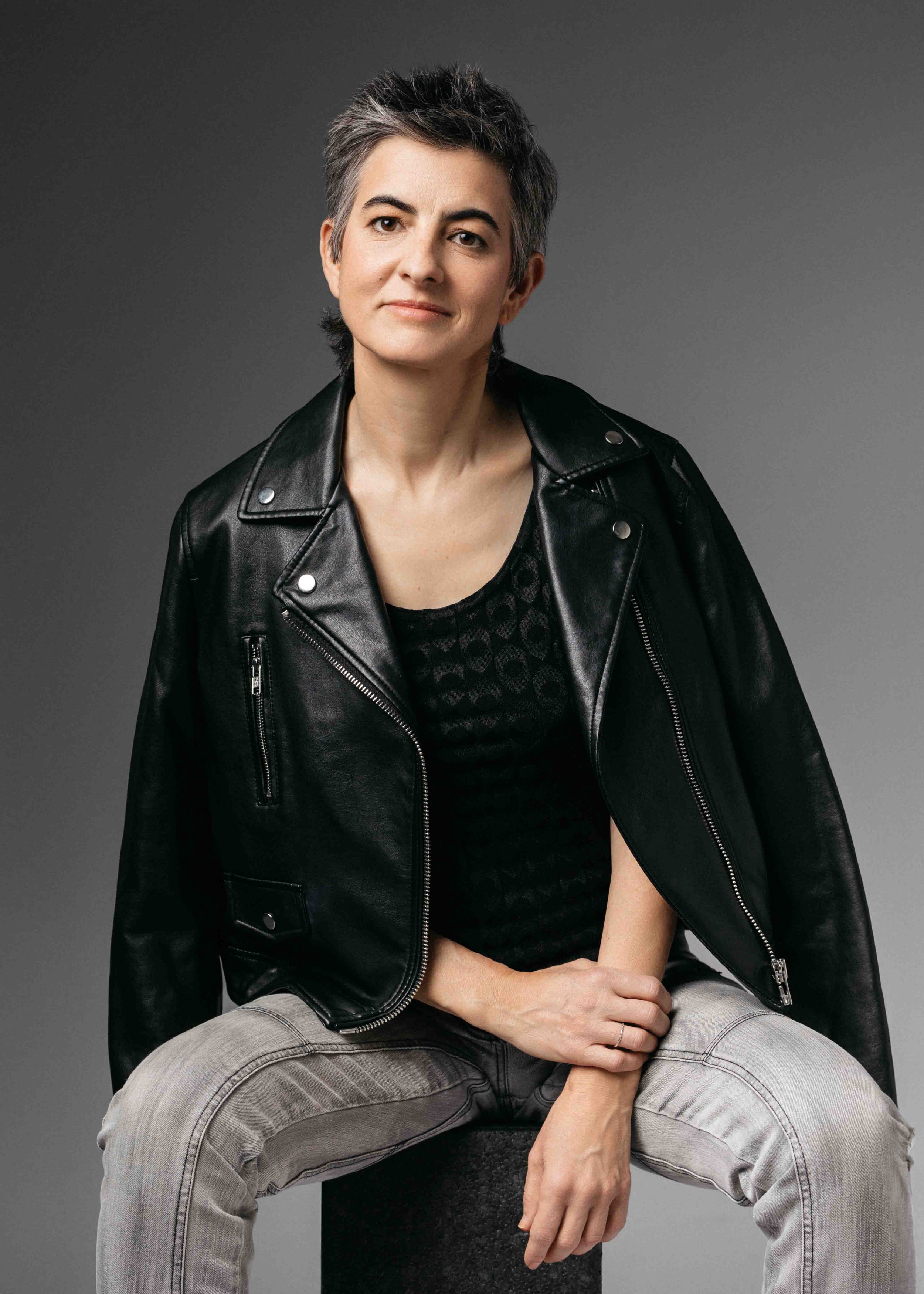 A woman with short gray hair wearing a black leather jacket and gray pants, sitting on a stool against a plain gray background.