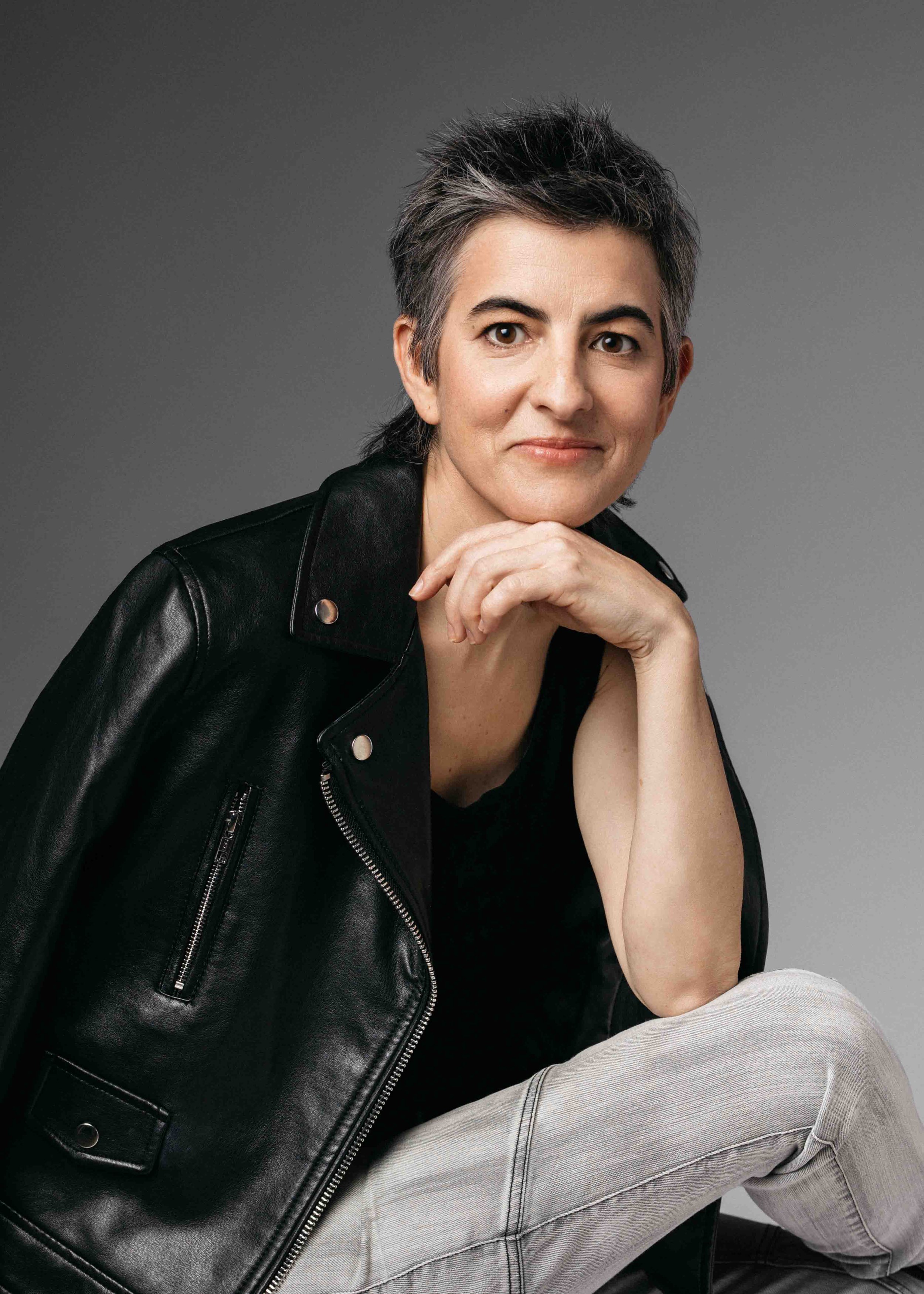 Portrait of a woman with short, gray hair, sitting with her hand resting on her chin, wearing a black leather jacket and light-colored jeans, against a gray background.