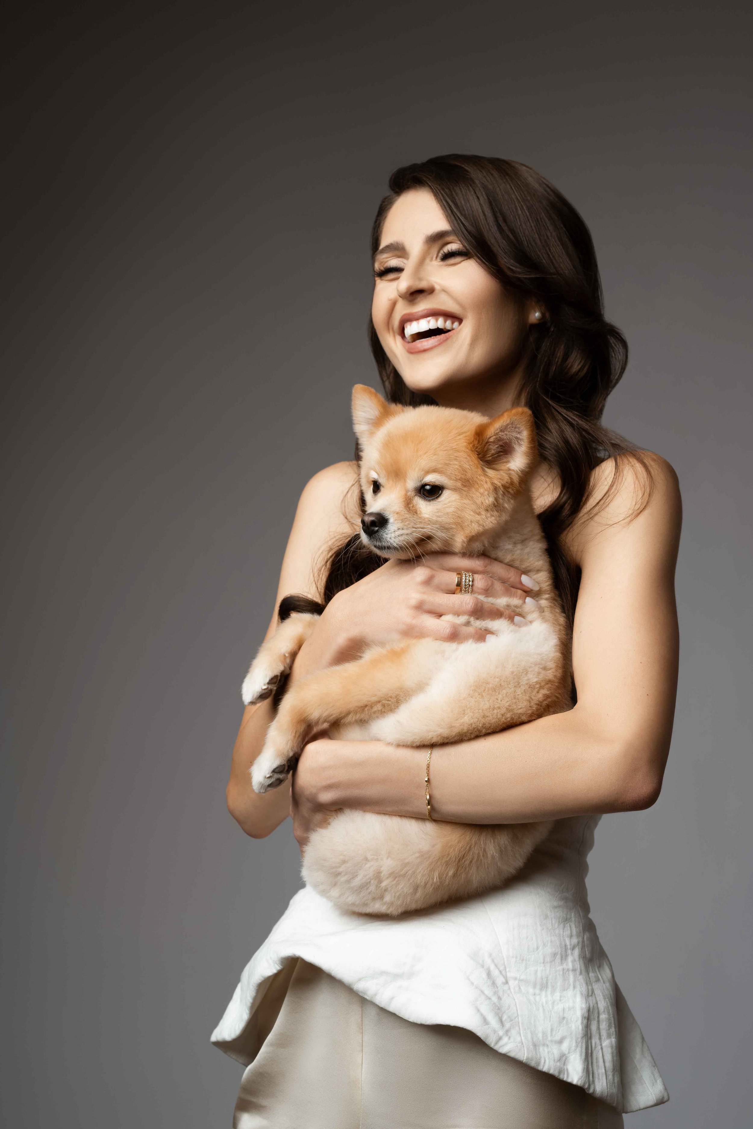 A woman with long dark hair, smiling and holding a small, fluffy puppy, against a plain gray background.