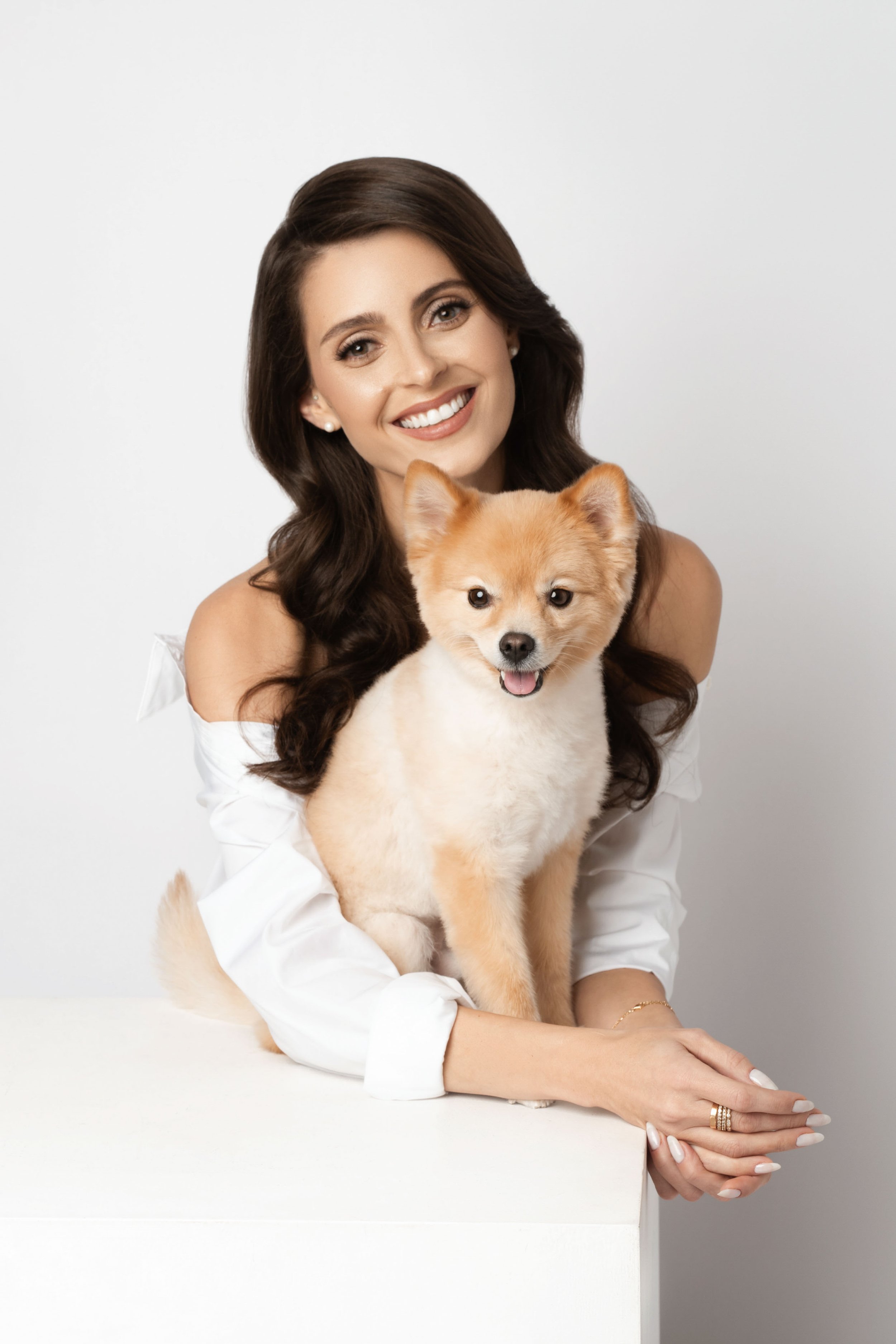 A smiling woman with long dark hair holding a small, fluffy, light-colored dog against a plain white background.