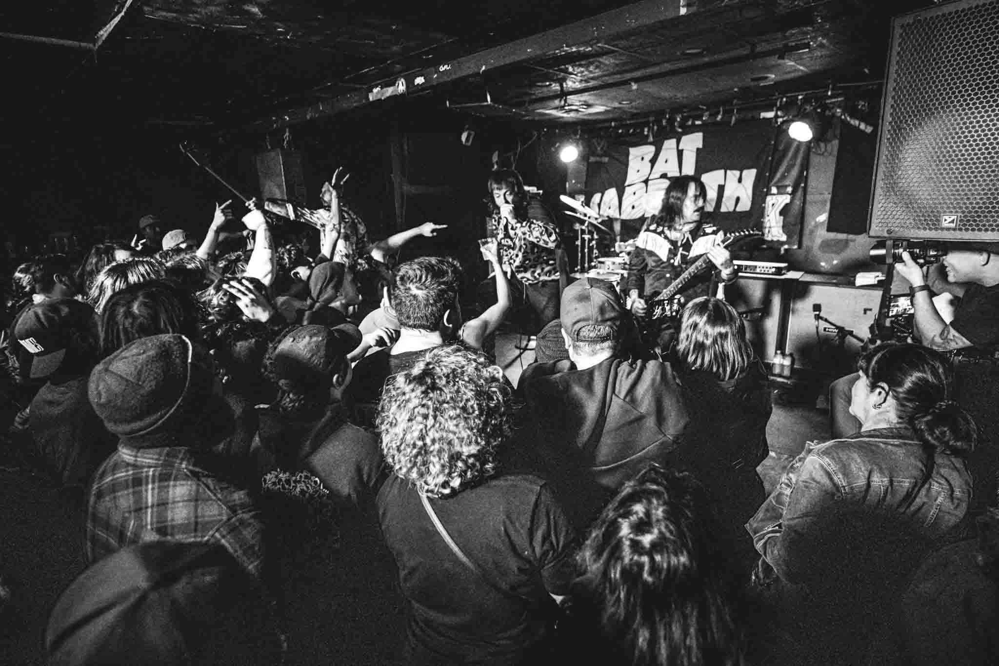 Black and white photo of a live music performance at a small venue with three female musicians on stage and audience members watching and dancing.
