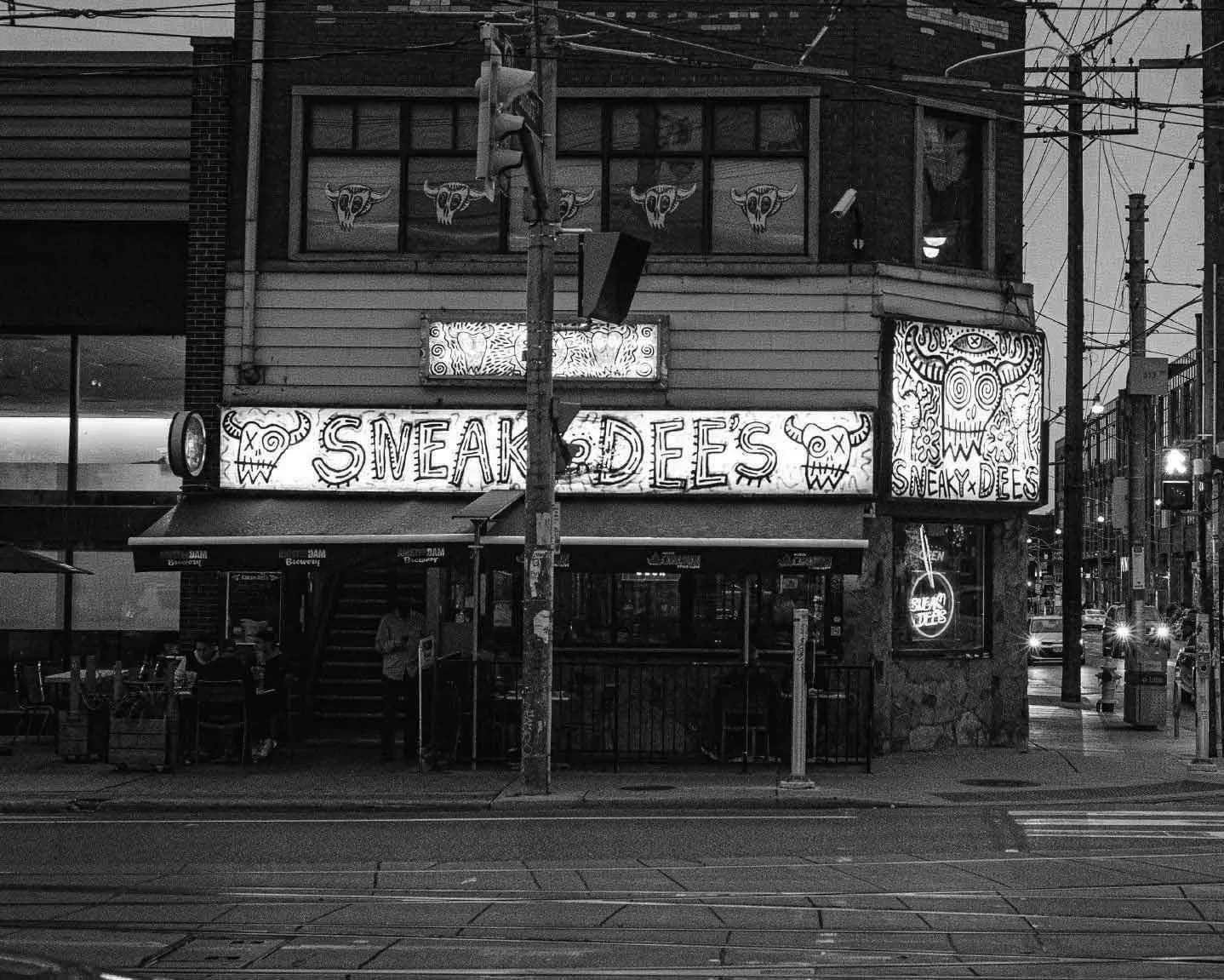 Street view of a two-story building with graffiti-style signage that reads 'Sneaky Dees,' featuring skull and horn illustrations. The building has illuminated signs, one on the lower right side and another with the establishment's name, and is located on a city street with visible tram tracks, utility poles, and parked cars.