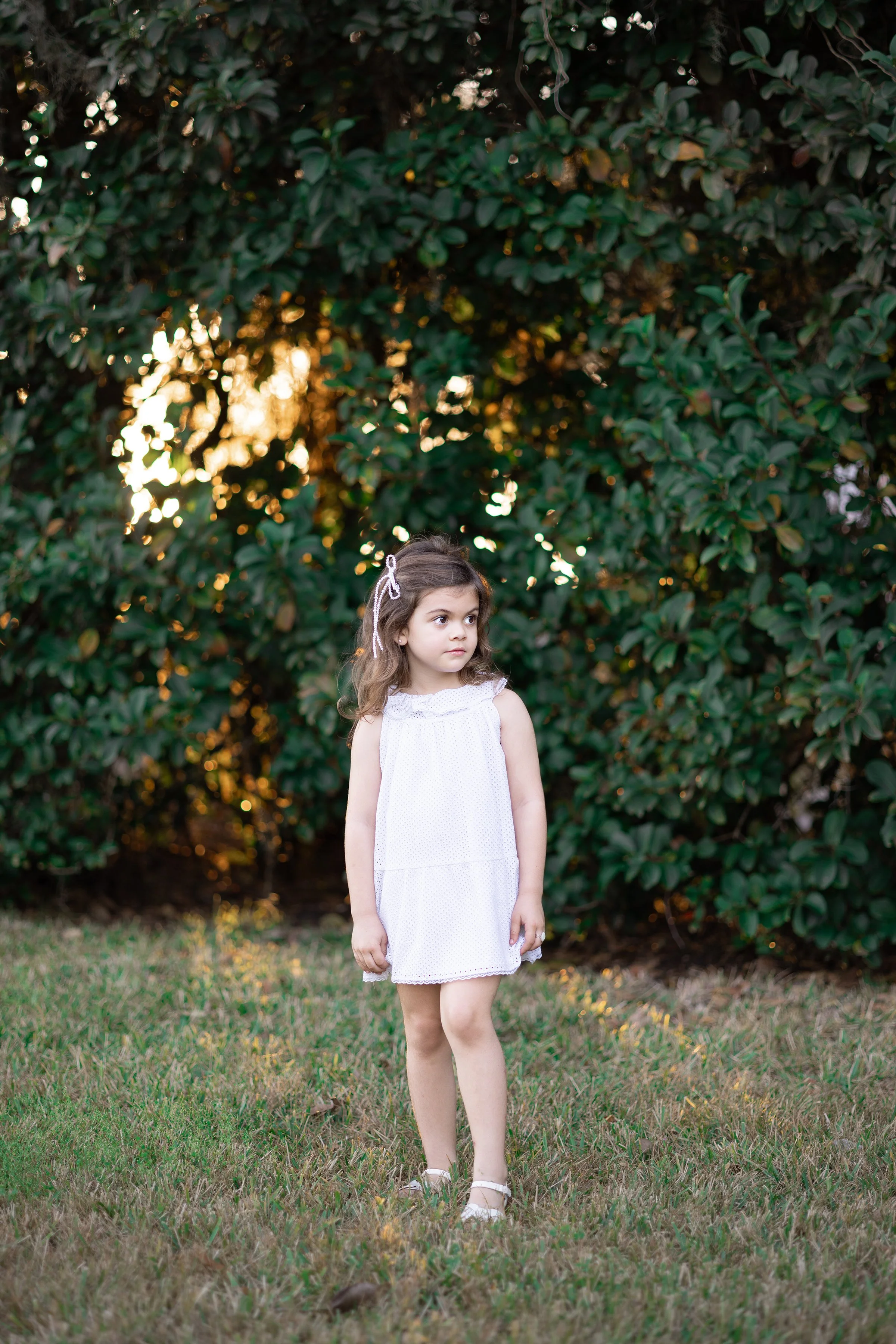 Young girl with long brown hair, wearing a white dress and sandals, standing on grass in front of a large bush with sunlight filtering through.
