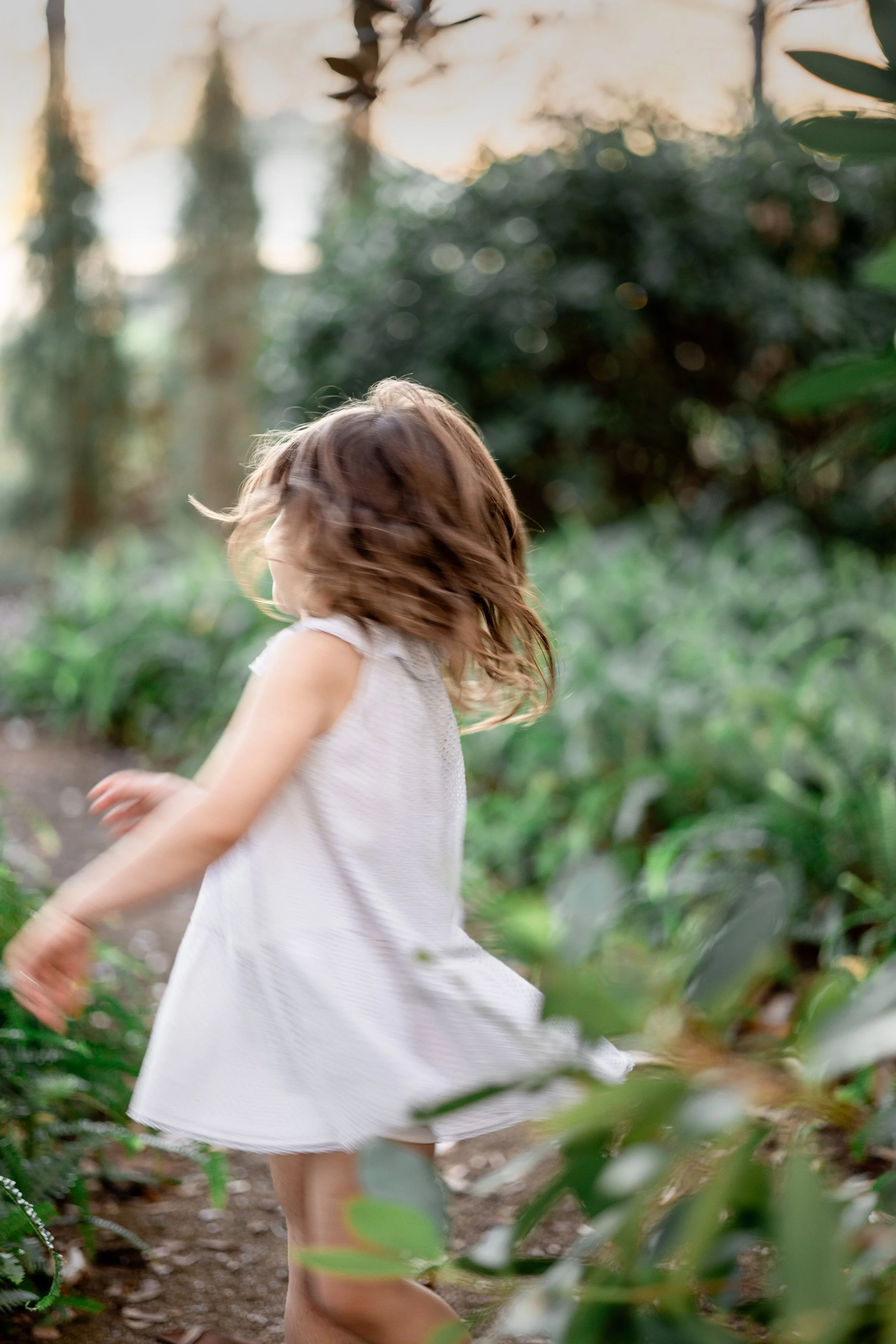 A young girl with brown curly hair spinning in a green outdoor garden or park during sunset, wearing a white dress.