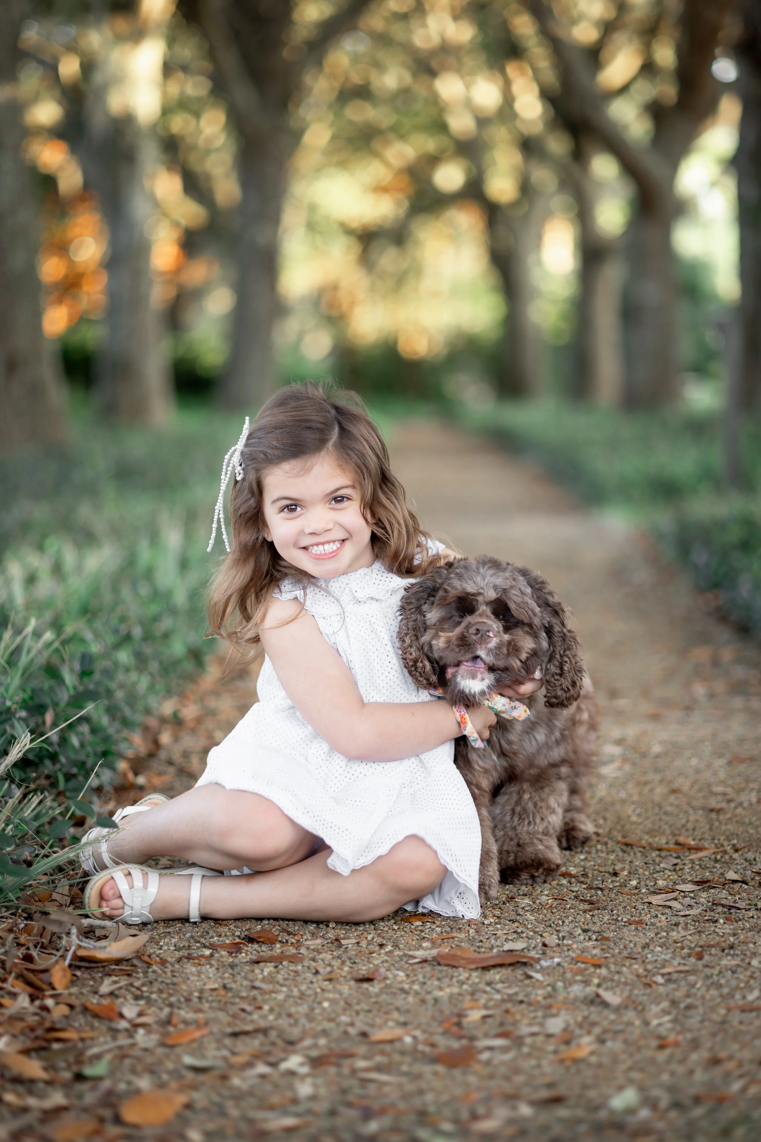 A young girl with brown hair and a white dress smiling while holding a brown, curly-haired puppy on a wooded path.