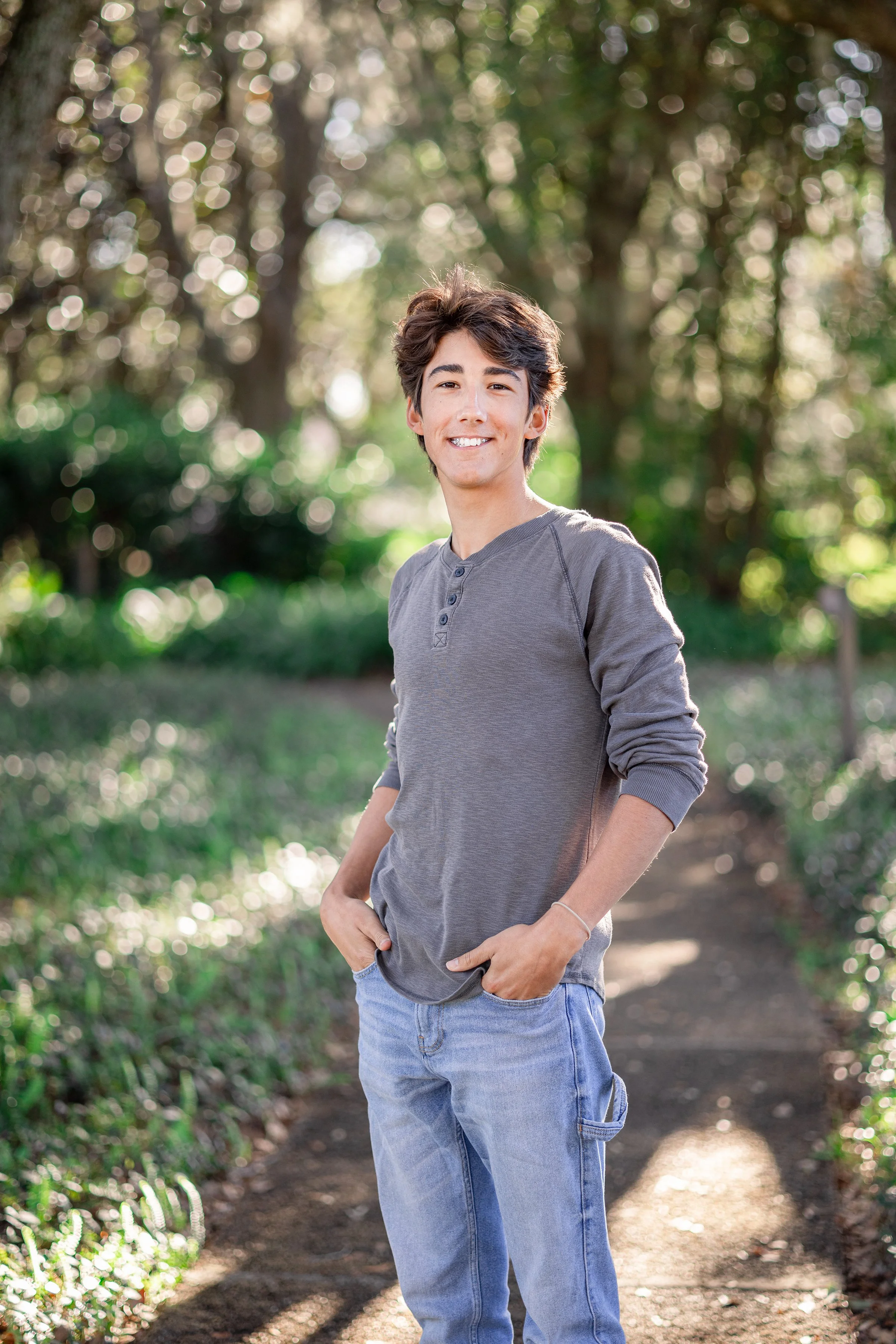 A young man with dark hair smiling, standing on a dirt path in a wooded area with sunlight filtering through the trees.