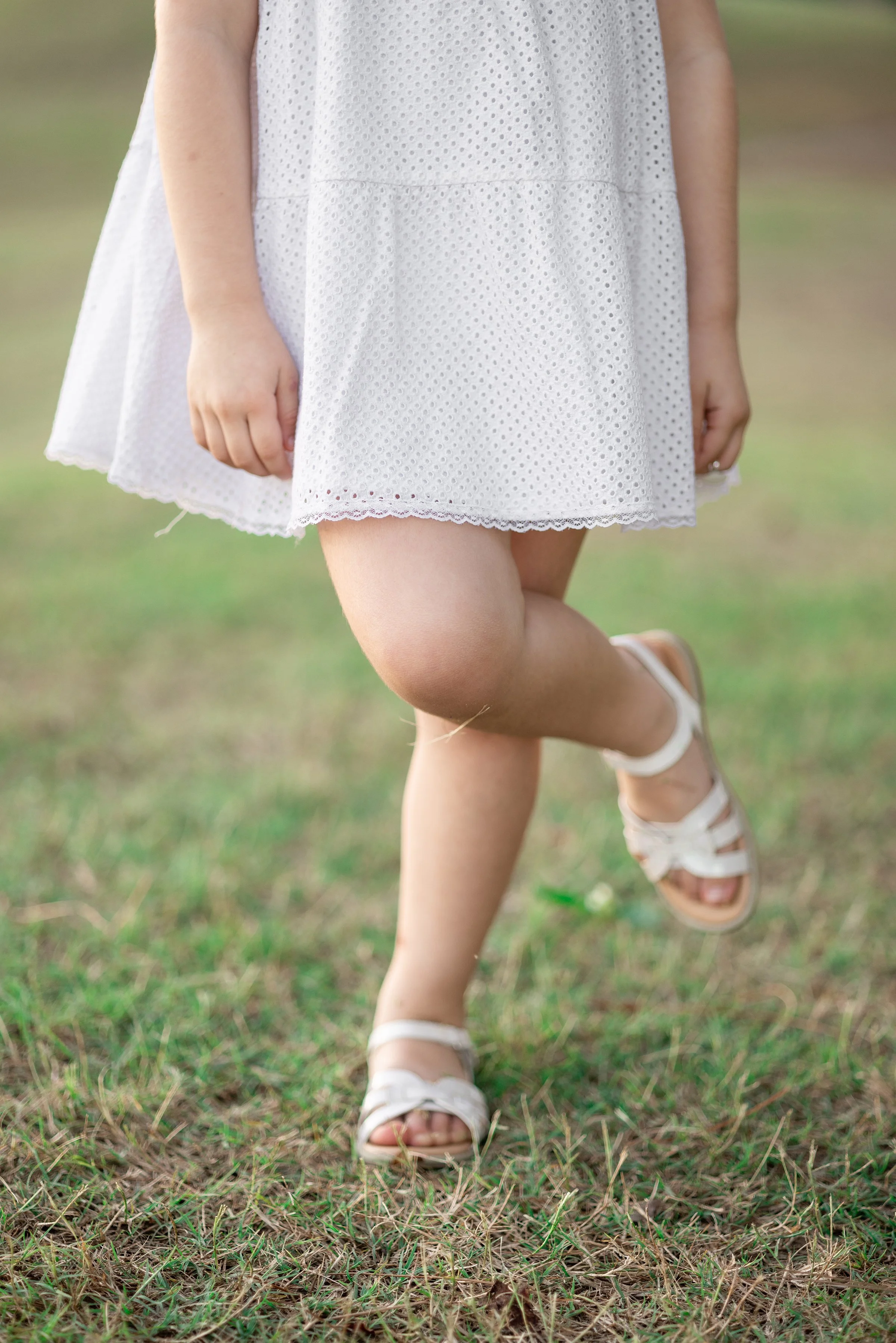 Child wearing a white dress and white sandals standing on grass, with one leg lifted.