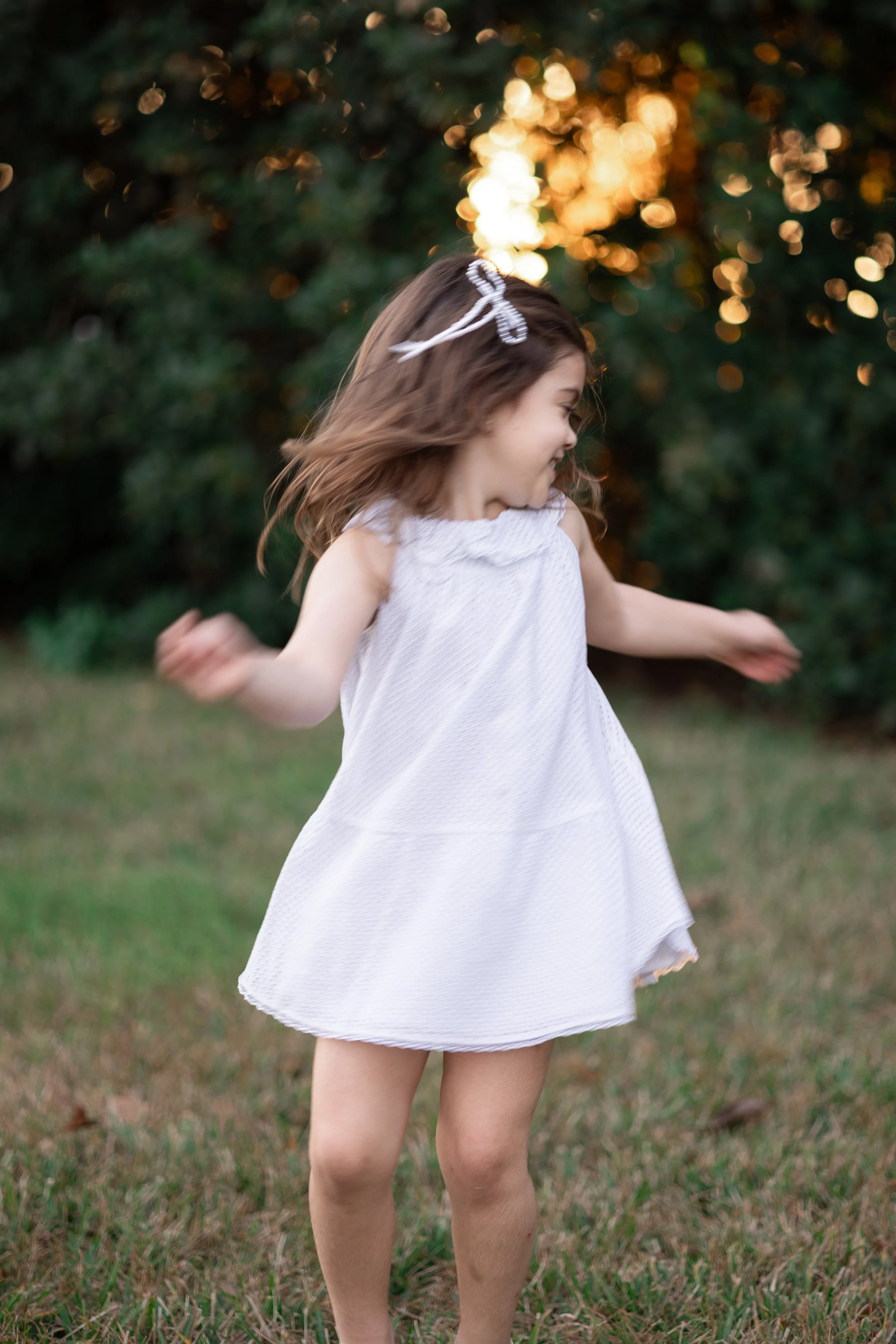 Young girl in a white dress spinning and smiling outside during golden hour with trees and sunlight in the background.