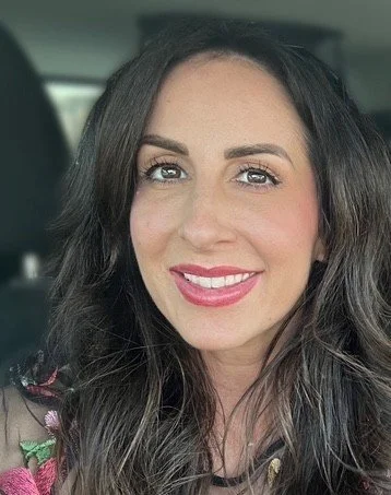 A woman with long dark hair, light skin, and makeup, smiling inside a car.