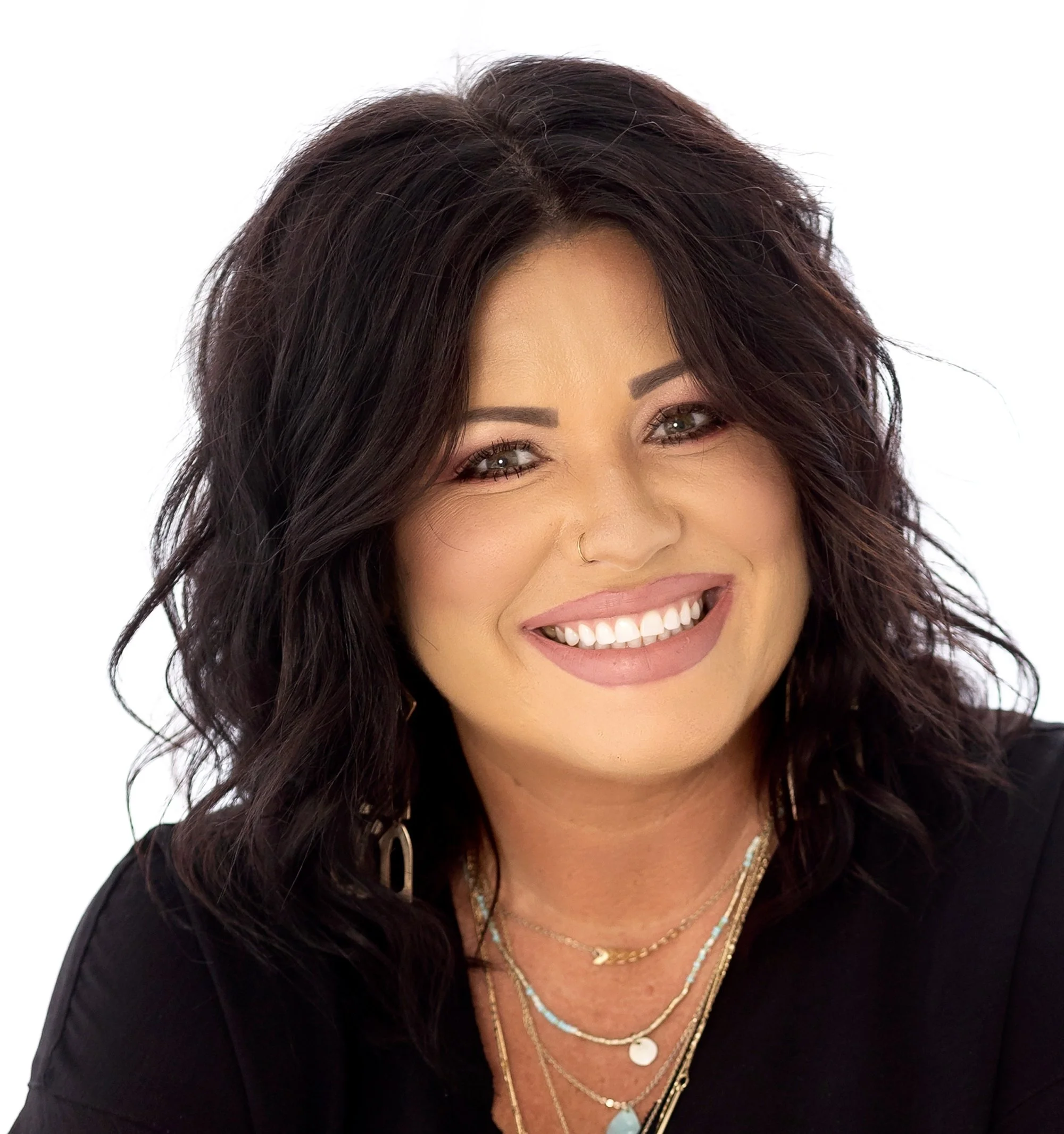 Close-up photo of a woman with dark brown, wavy hair, smiling and showing her teeth. She is wearing a black top and multiple layered necklaces, including a gold chain, turquoise beads, and a shell pendant. The background is plain white.