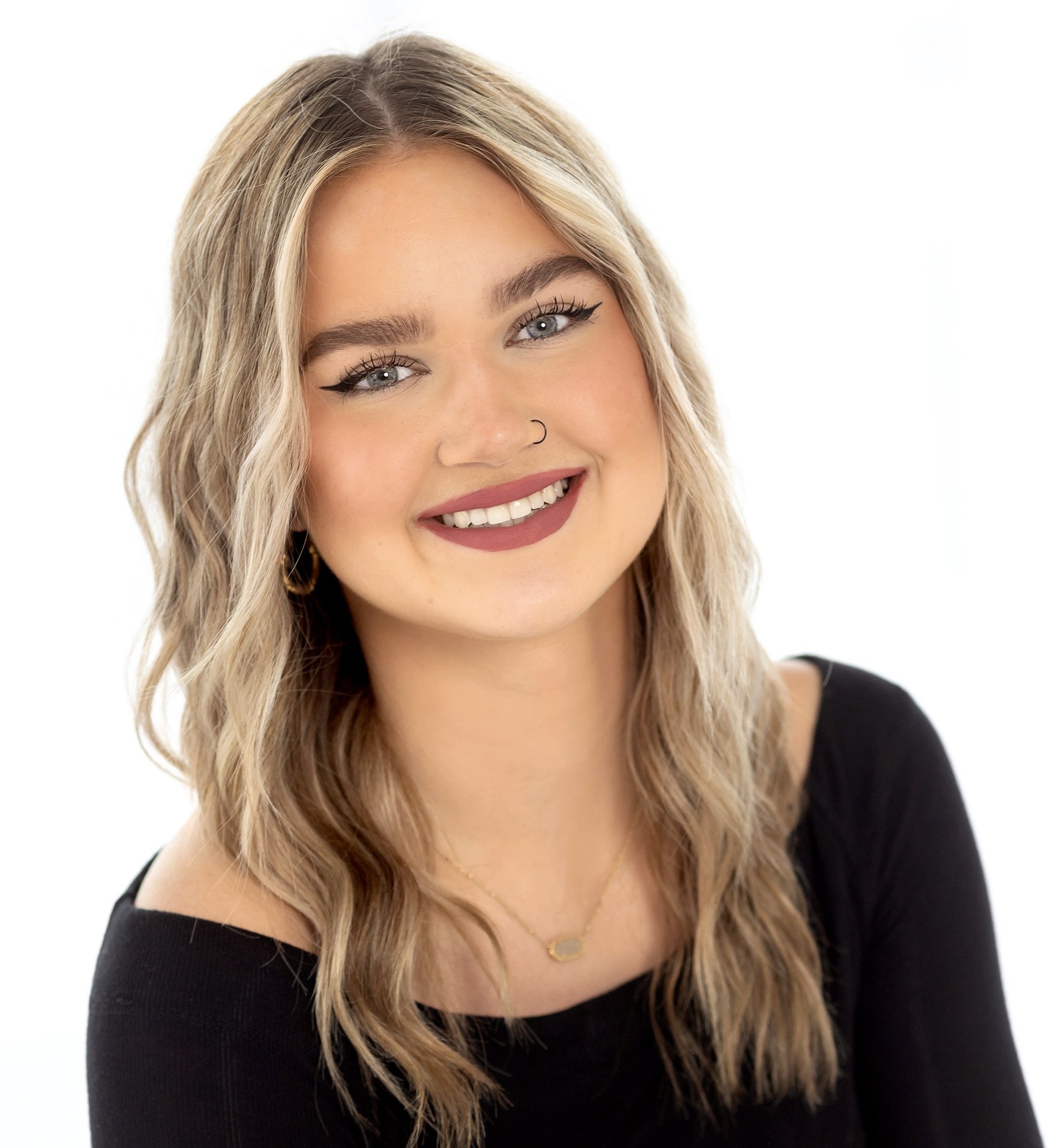 A young woman with blonde hair and blue eyes smiling, wearing a black top, gold necklace, and a nose ring, against a white background.