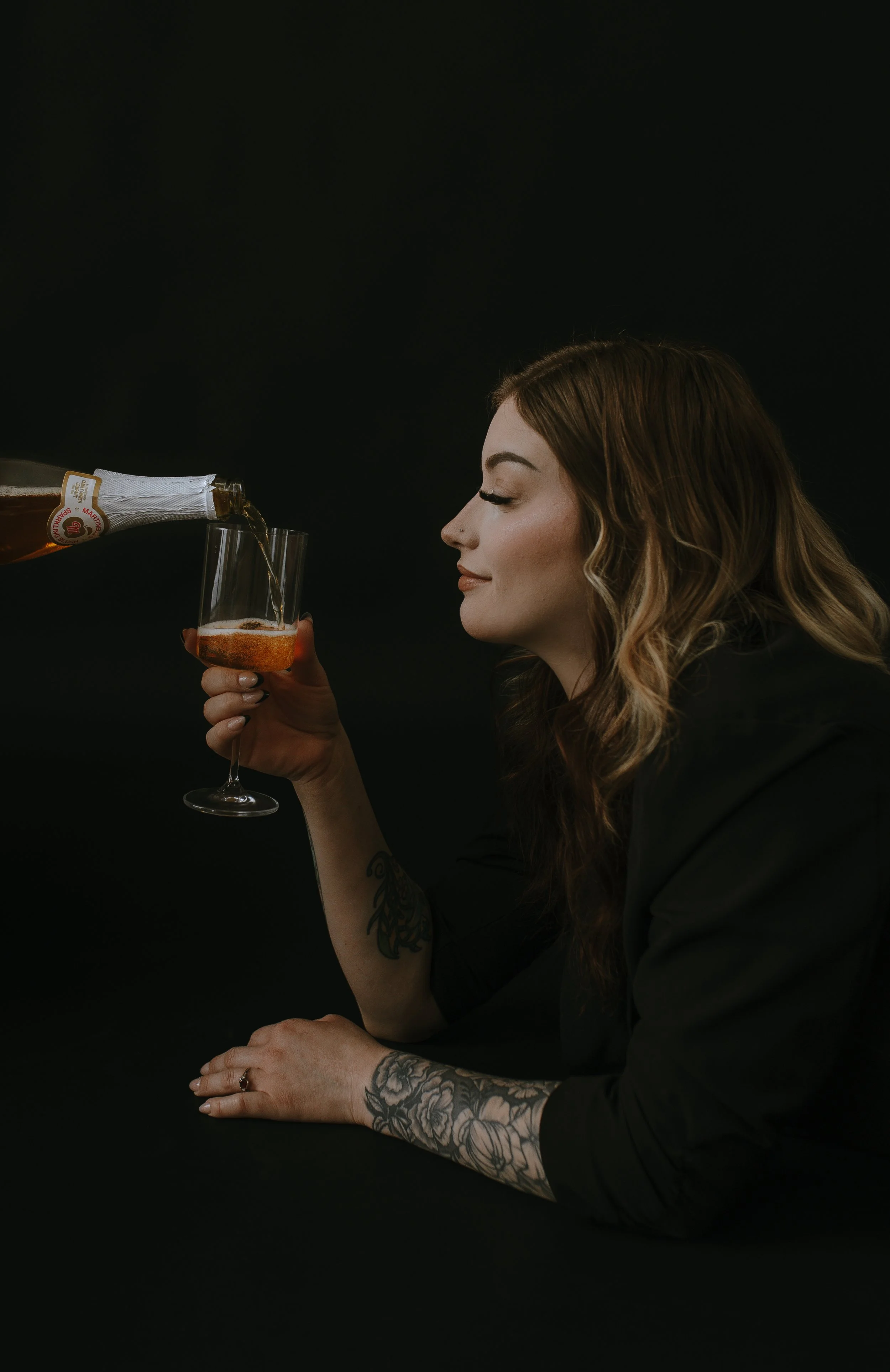 A woman with long wavy hair, tattoos on her arm, and a nose piercing is sitting against a black background, holding a wine glass with a dark liquid while a bottle is pouring another dark liquid into her glass.