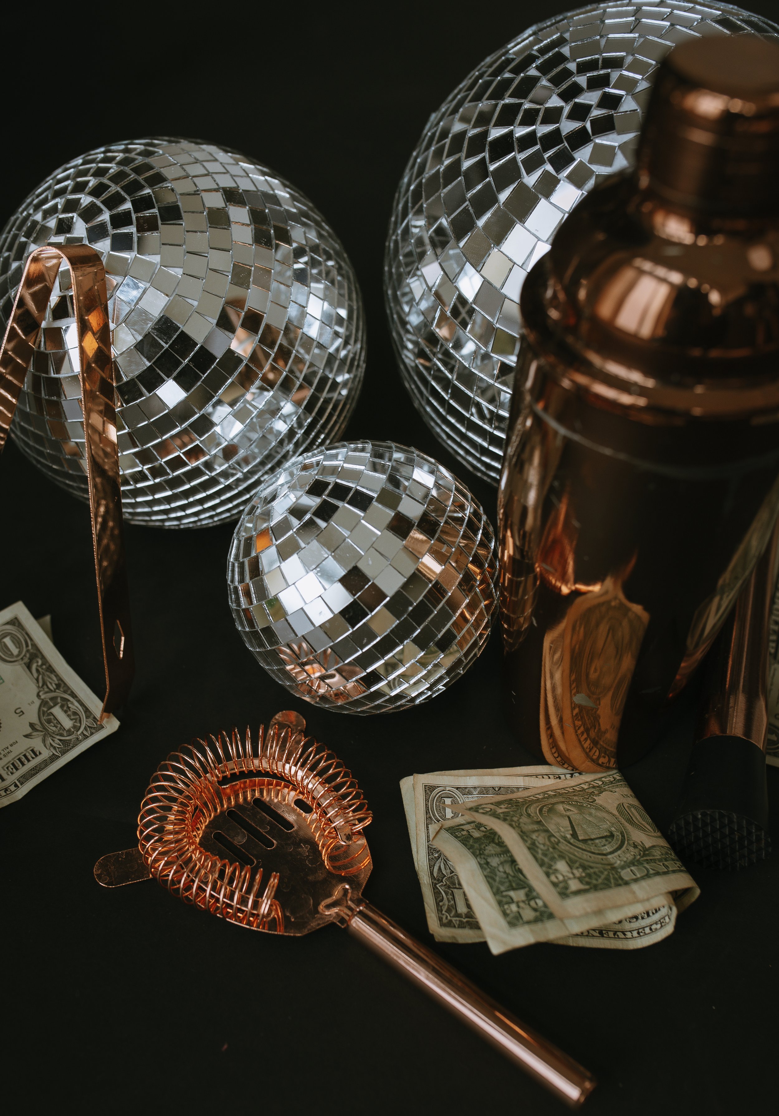 Disco balls, copper-colored stirrers, and folded dollar bills on a black surface.