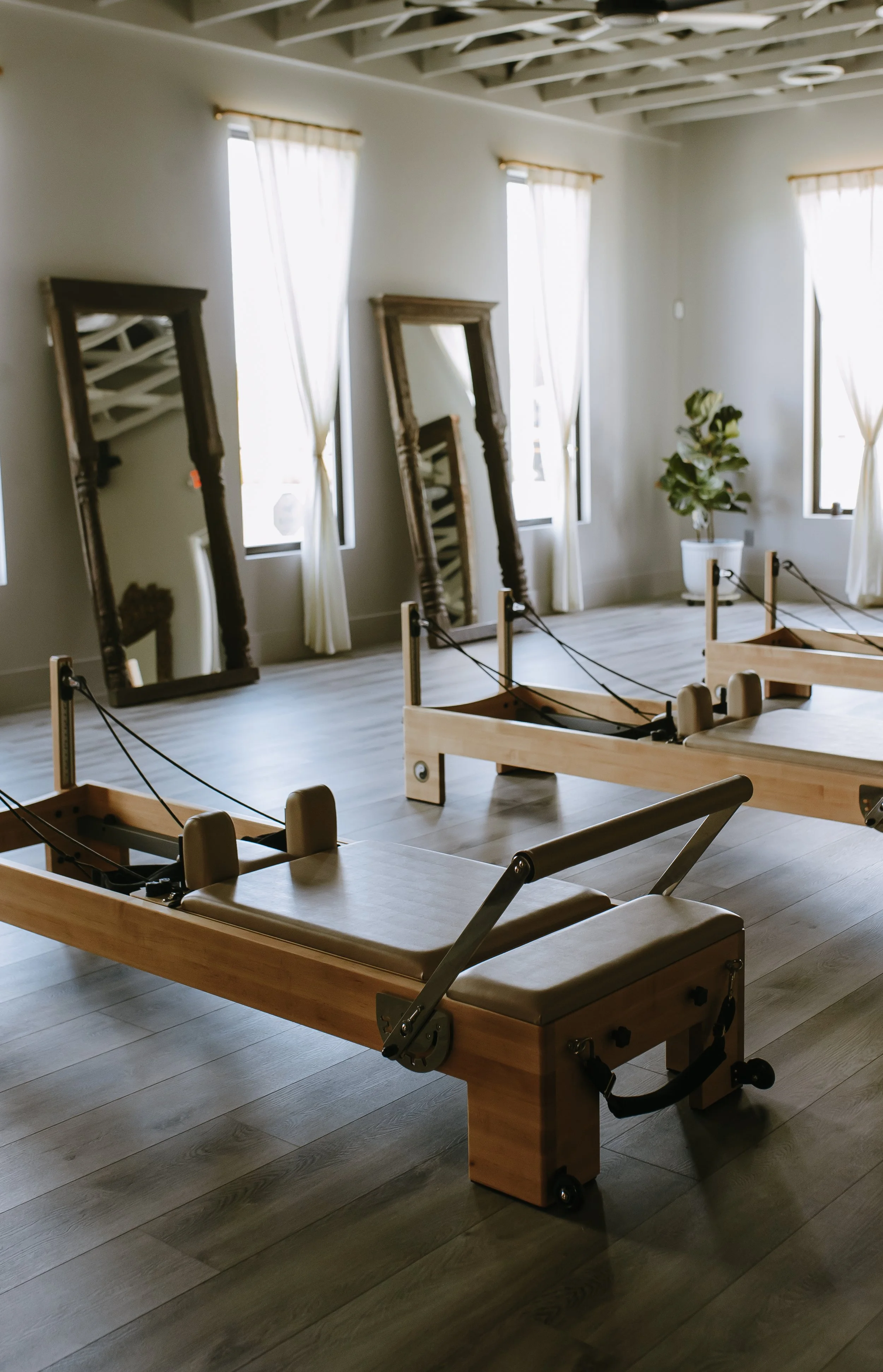 A wooden therapy or examination table in a room with light-colored walls, three tall mirrors leaning against the wall, a potted plant, and three windows with white curtains letting in natural light.