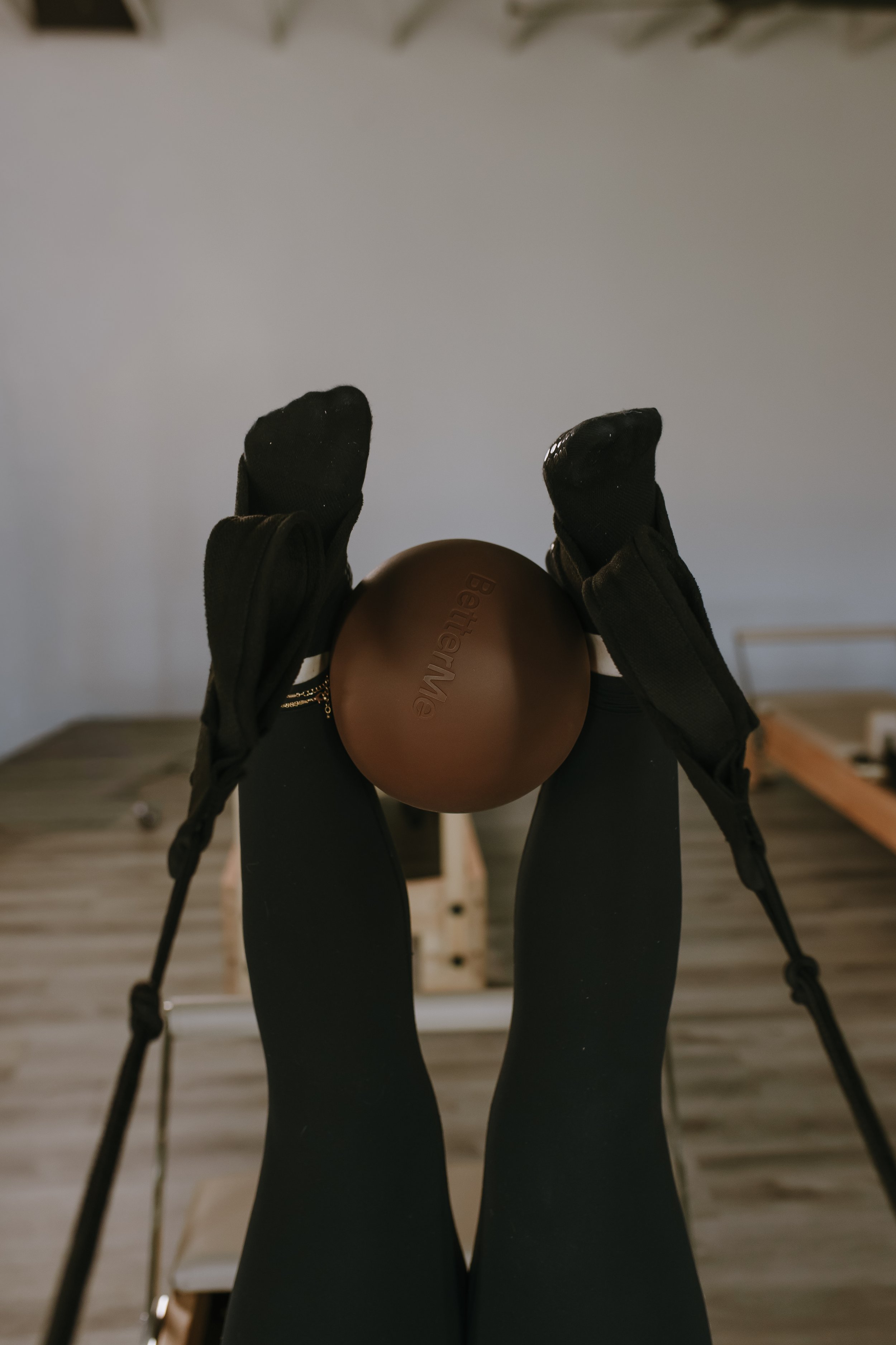 Person wearing black gloves and black leggings holding a brown exercise ball with 'Bette' written on it, standing on a wooden floor in a room with workout equipment and a white wall.