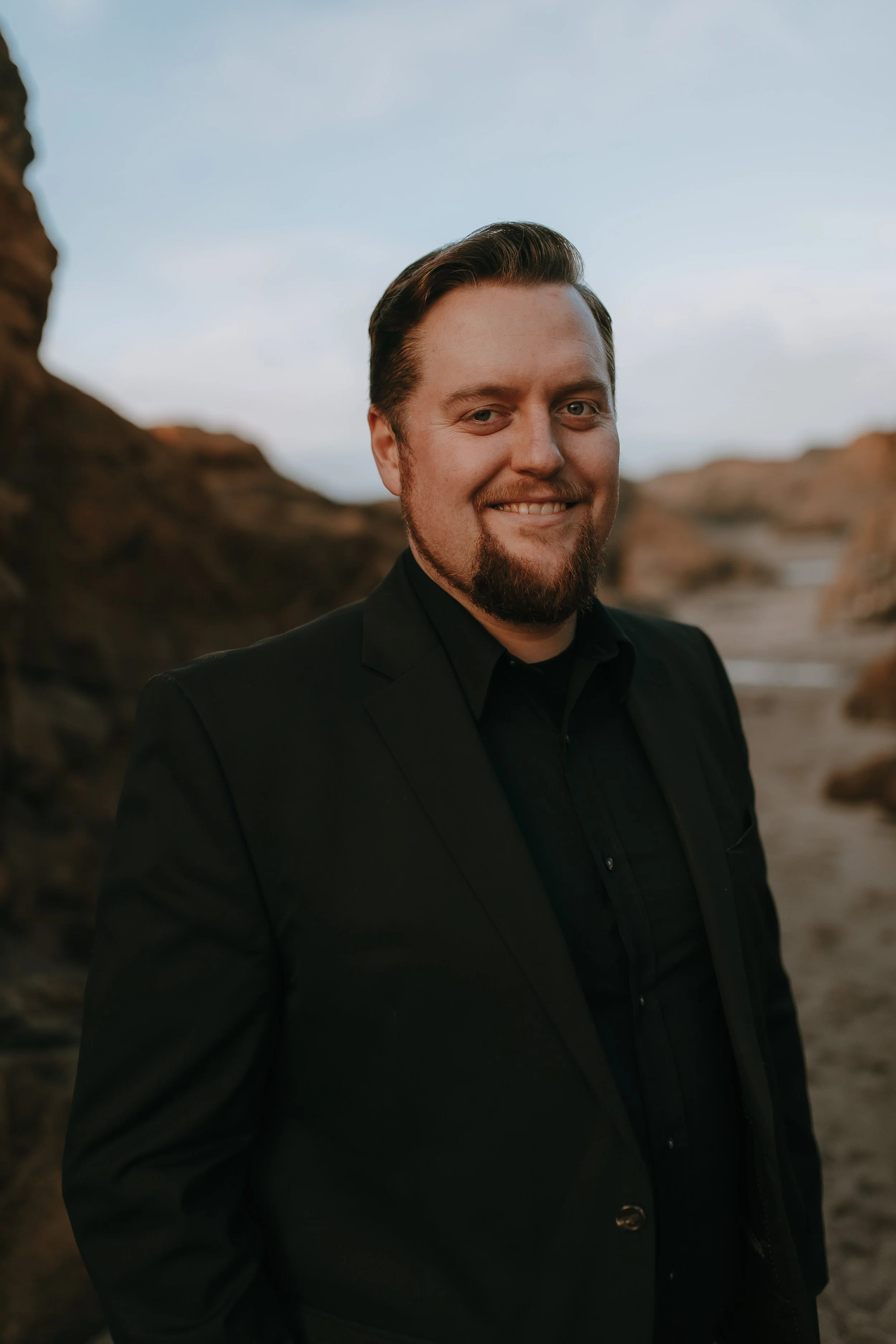 A man with a beard and short hair, wearing a black suit, smiling outdoors with rocky formations in the background.