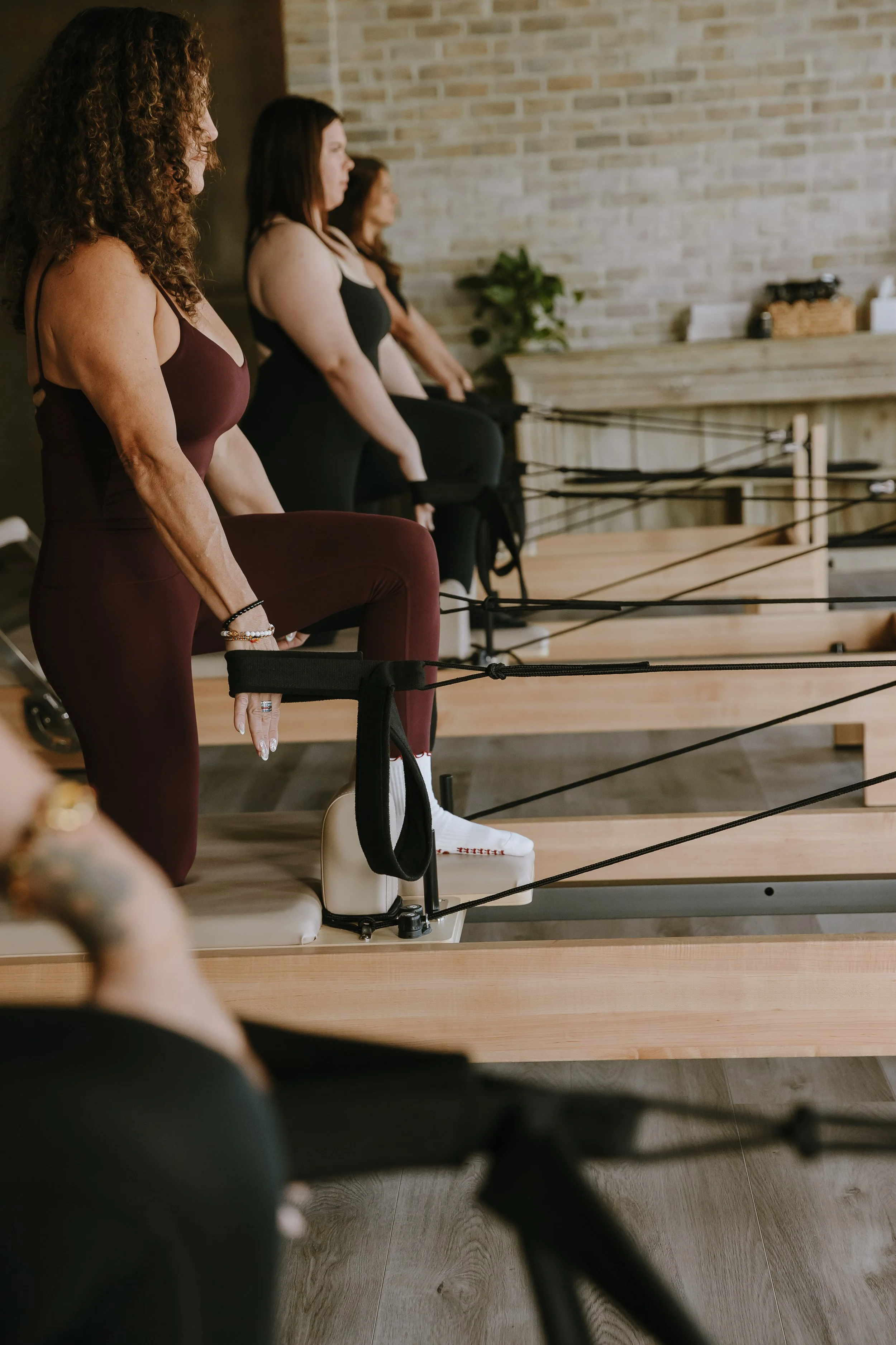Side view of three women in yoga class, kneeling in a pose with hands resting on yoga blocks, inside a room with brick wall and wooden flooring.