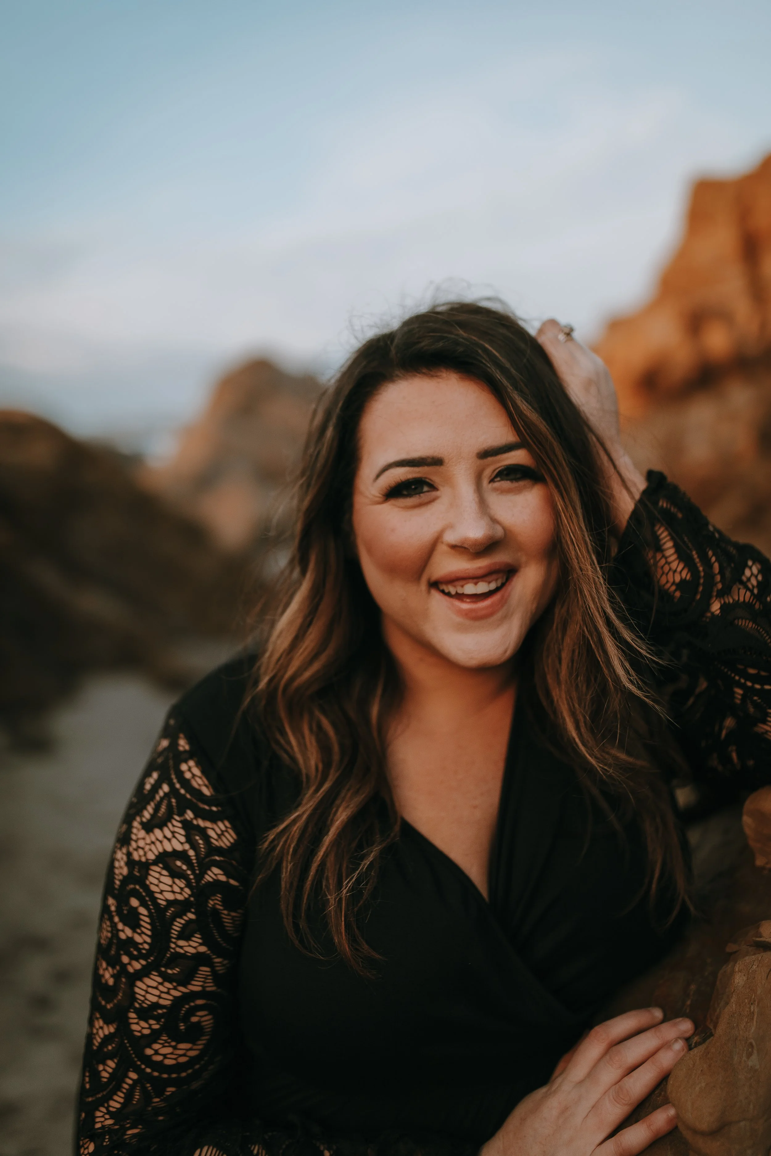 A woman smiling outdoors at sunset, with long wavy brown hair, wearing a black lace top, standing near rocks with a cloudy sky in the background.