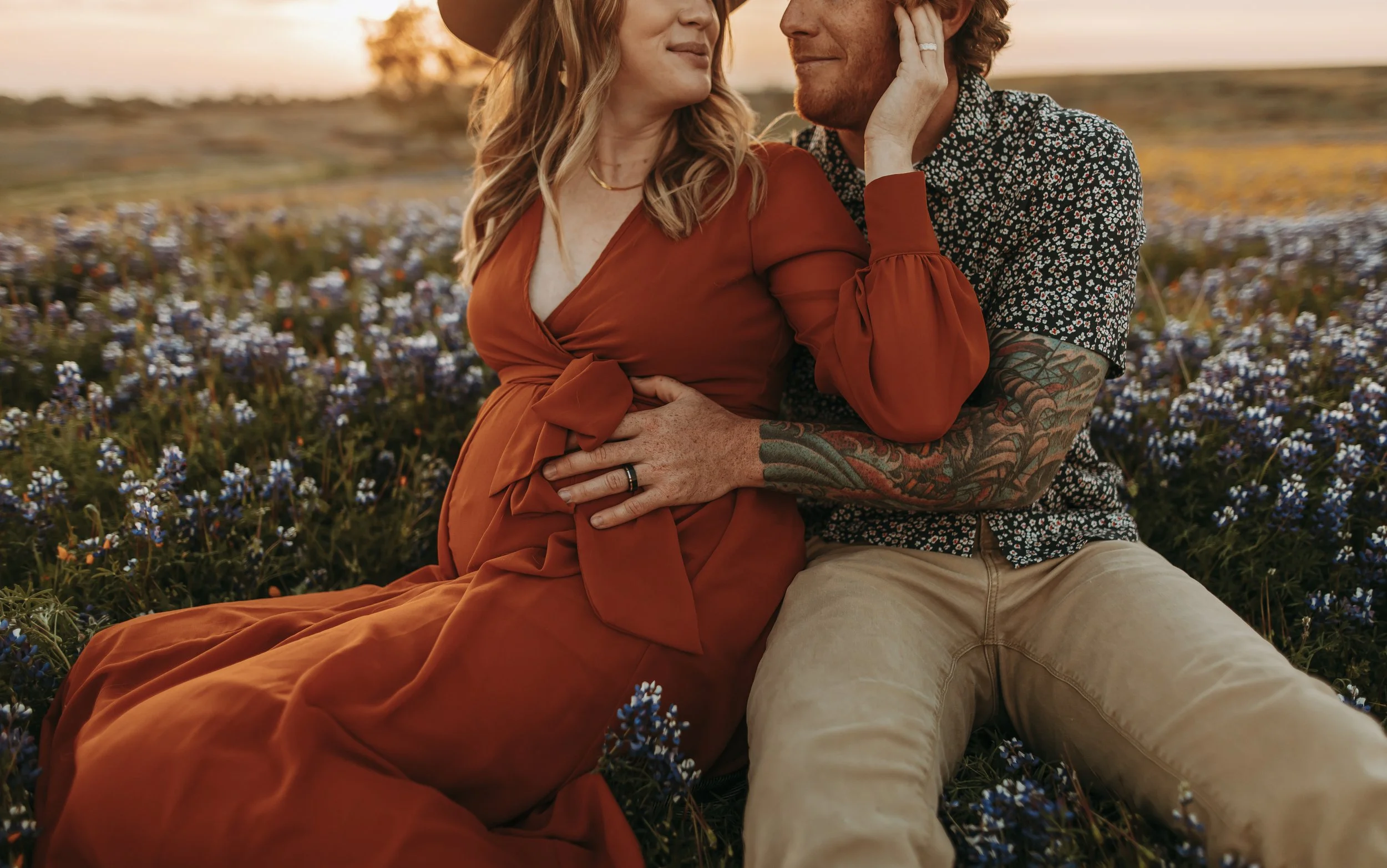 A couple sitting in a field of bluebonnet flowers during sunset, with the woman wearing a rust-colored dress and the man in a floral shirt, tenderly touching each other's faces.