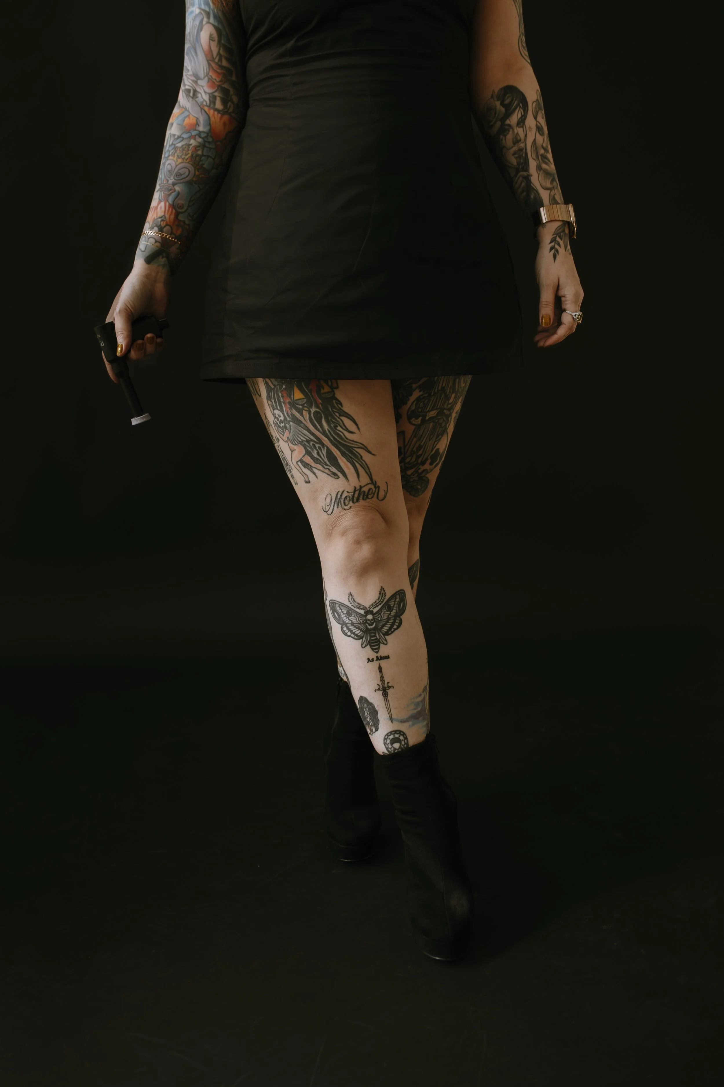 A person standing against a black background, wearing a black dress and black boots, holding a cigarette lighter in their right hand, with visible tattoos on their arms and legs including floral, face, and butterfly designs.