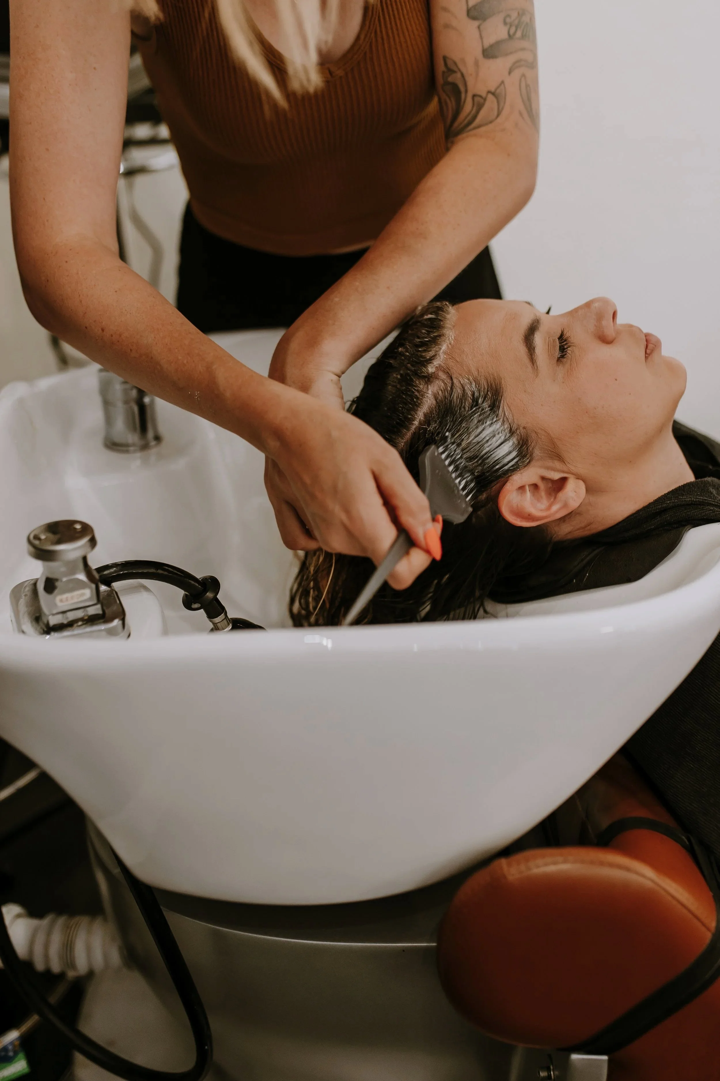 A woman is getting her hair washed in a salon sink, with her head tilted back into the white basin while a stylist rinses her hair.
