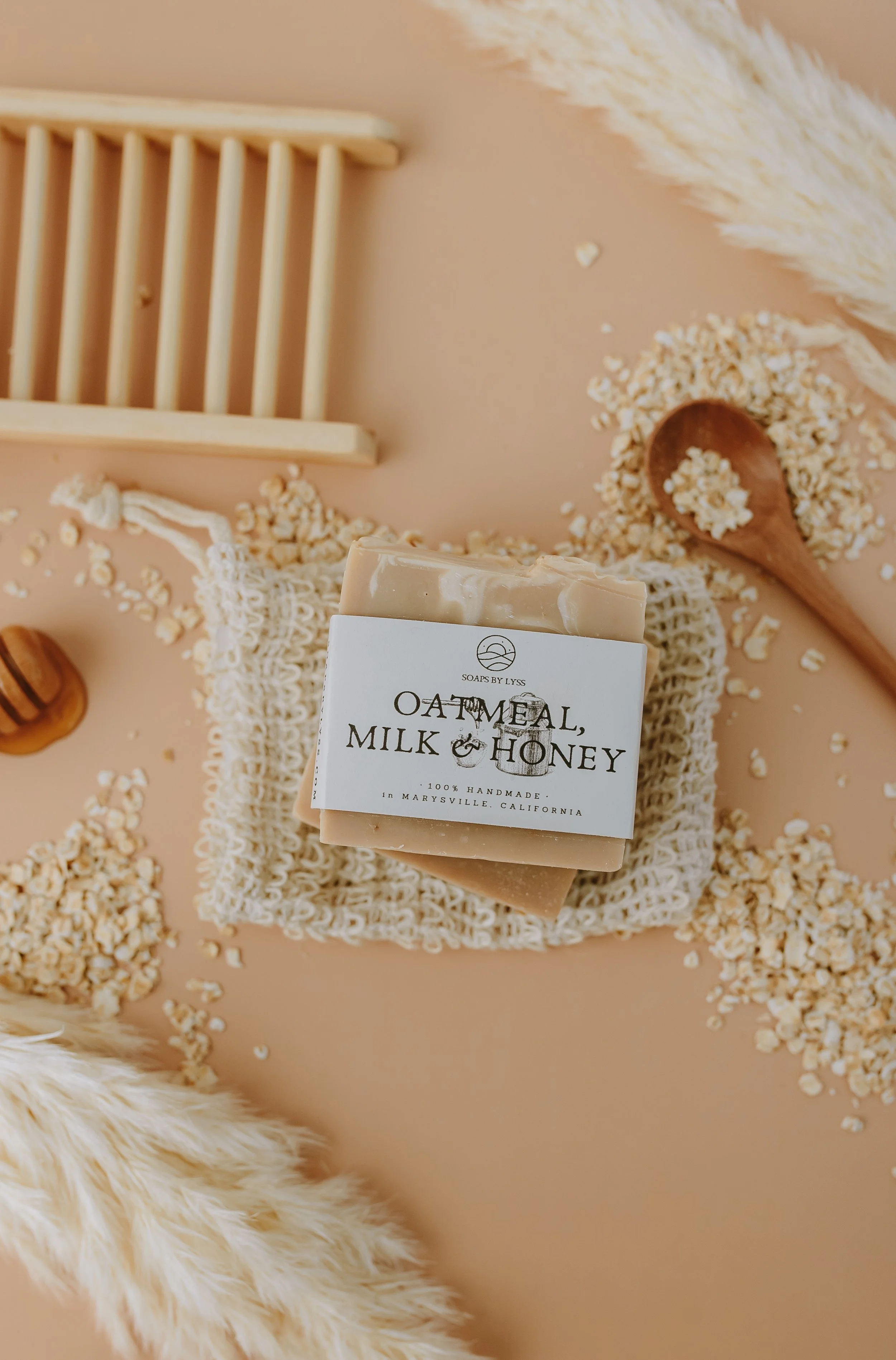 Bar of soap labeled "Oatmeal, Milk & Honey" resting on a textured cloth surrounded by scattered oatmeal flakes, with a wooden spoon filled with oatmeal flakes nearby, on a soft peach background with a white furry fabric in the corner.