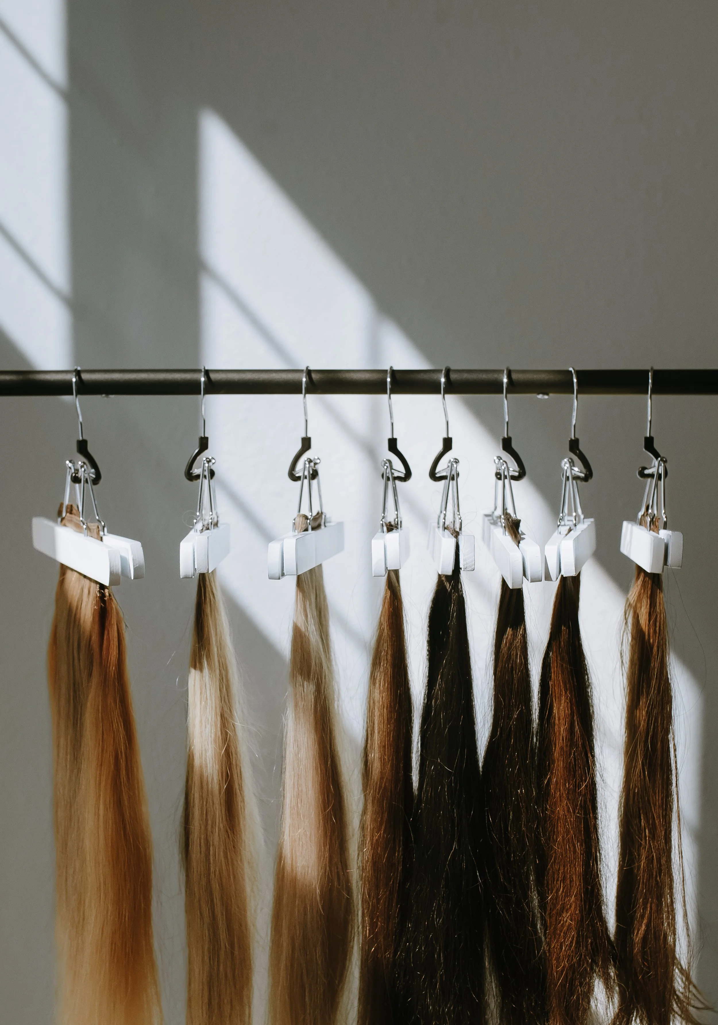 Multiple hair extensions hanging on a metal rod with clips, casting shadows on a white wall.