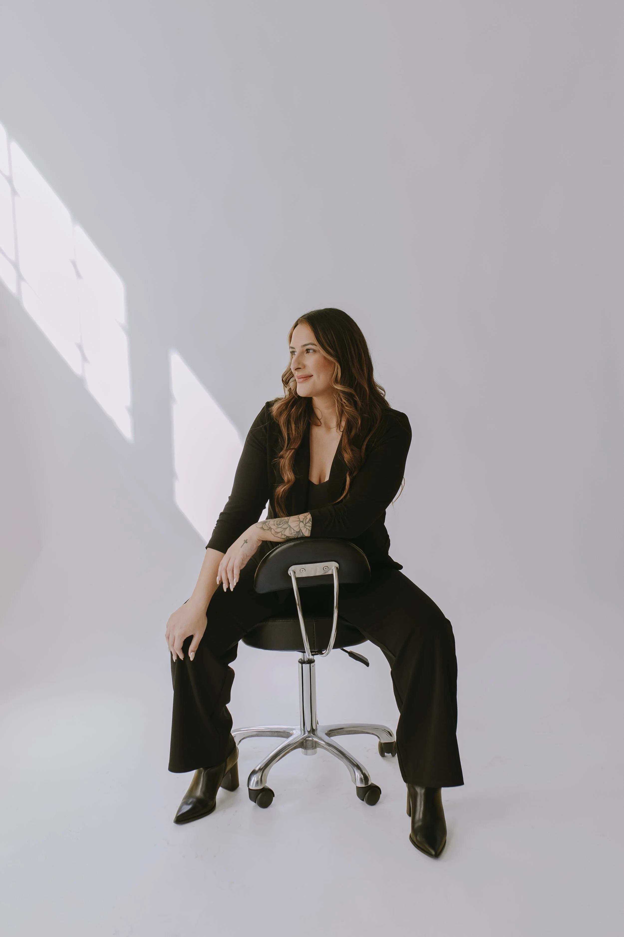 A woman with long brown hair, wearing a black outfit and black boots, sitting on a stool in a minimalistic studio with a white background and patterned sunlight shadows.