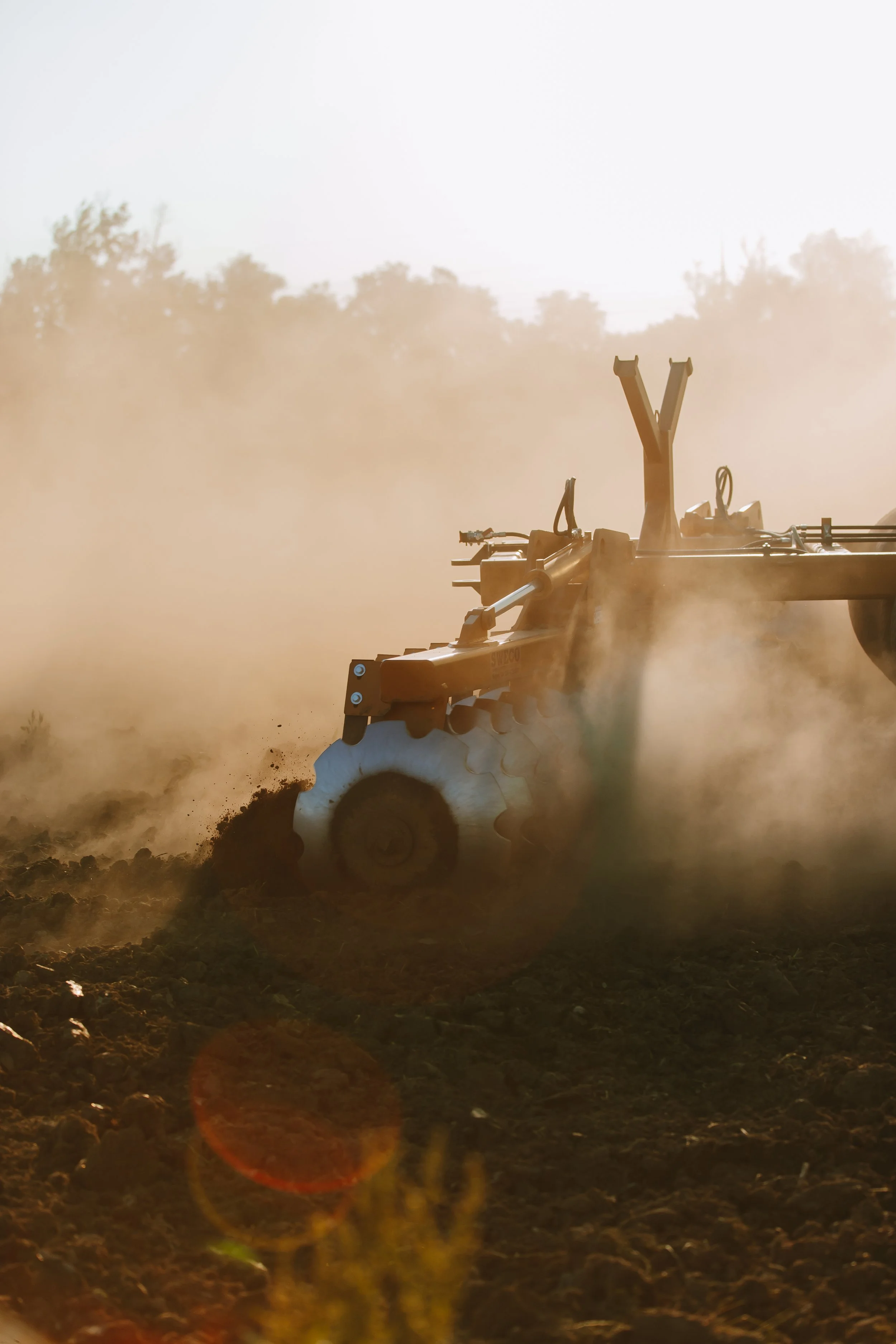 A tractor working on a farm with dust and soil in the air during sunset or sunrise.
